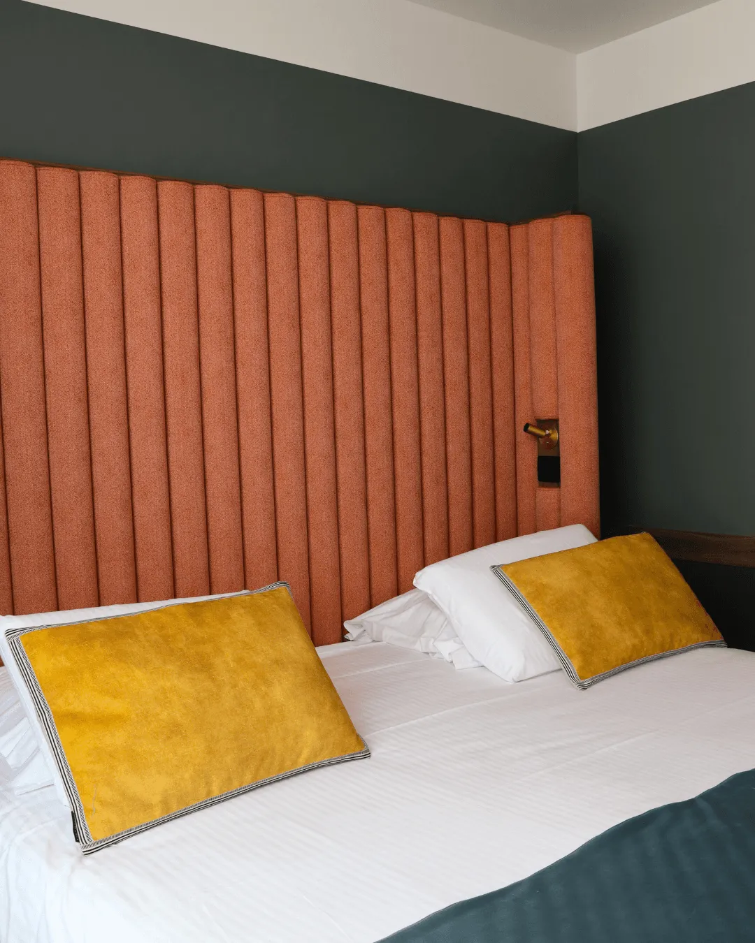 Lit avec une tête de lit orange, deux oreillers jaunes et des draps blancs dans une chambre aux murs sombres