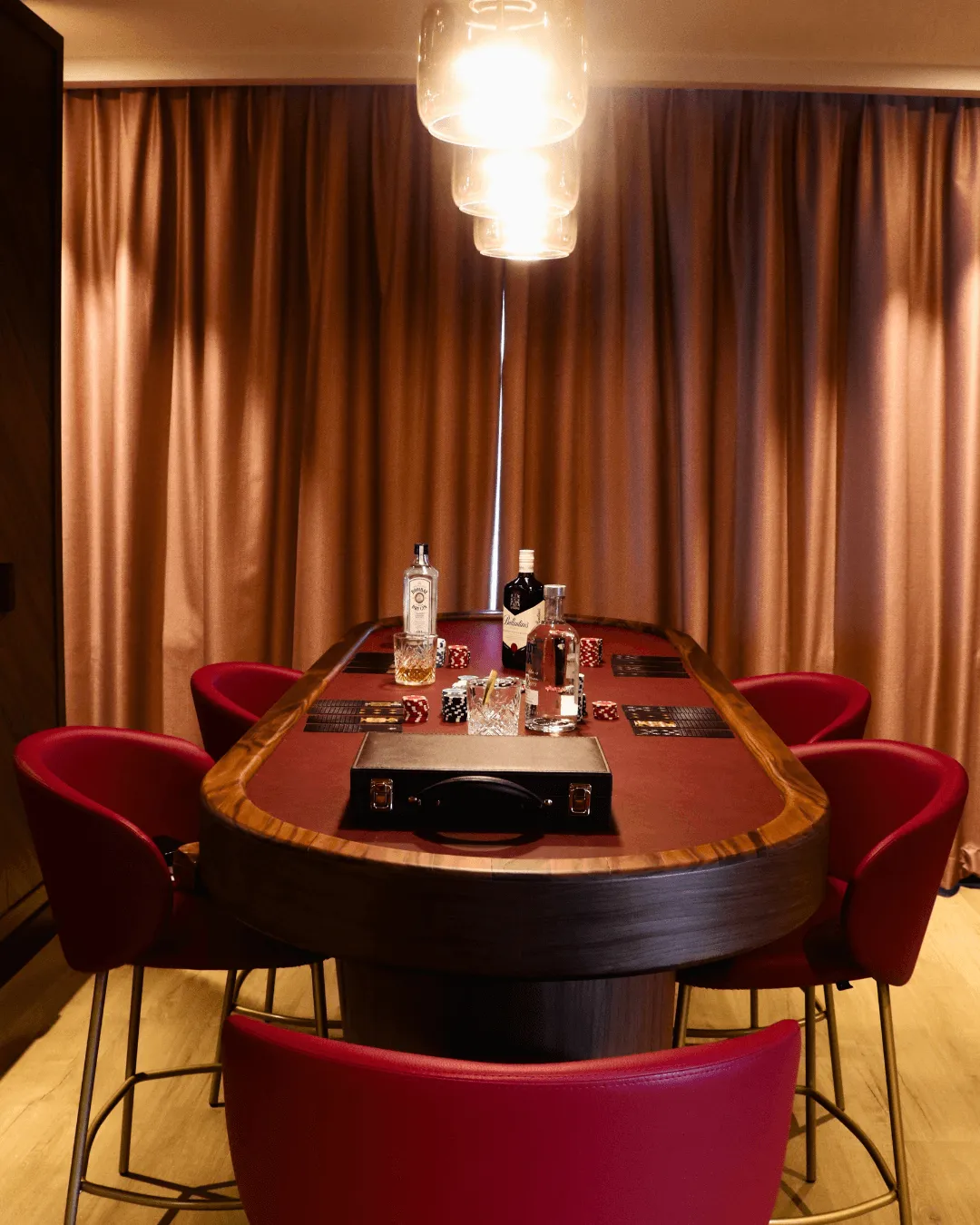 Table de poker en bois avec six chaises rouges, décorée de bouteilles, verres et jetons, éclairée par un luminaire suspendu dans une pièce aux rideaux dorés
