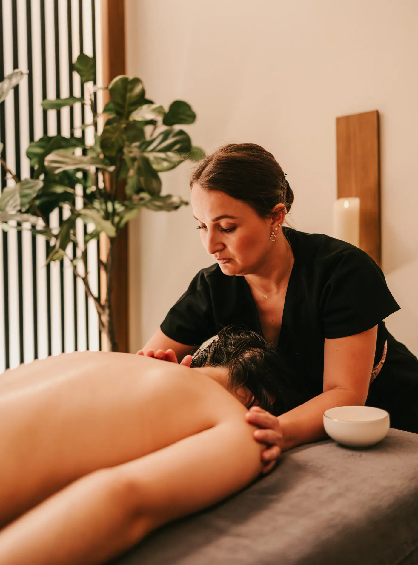 Une femme en noir donne un massage à une personne allongée sur le ventre dans un cadre intérieur avec une plante verte et une bougie