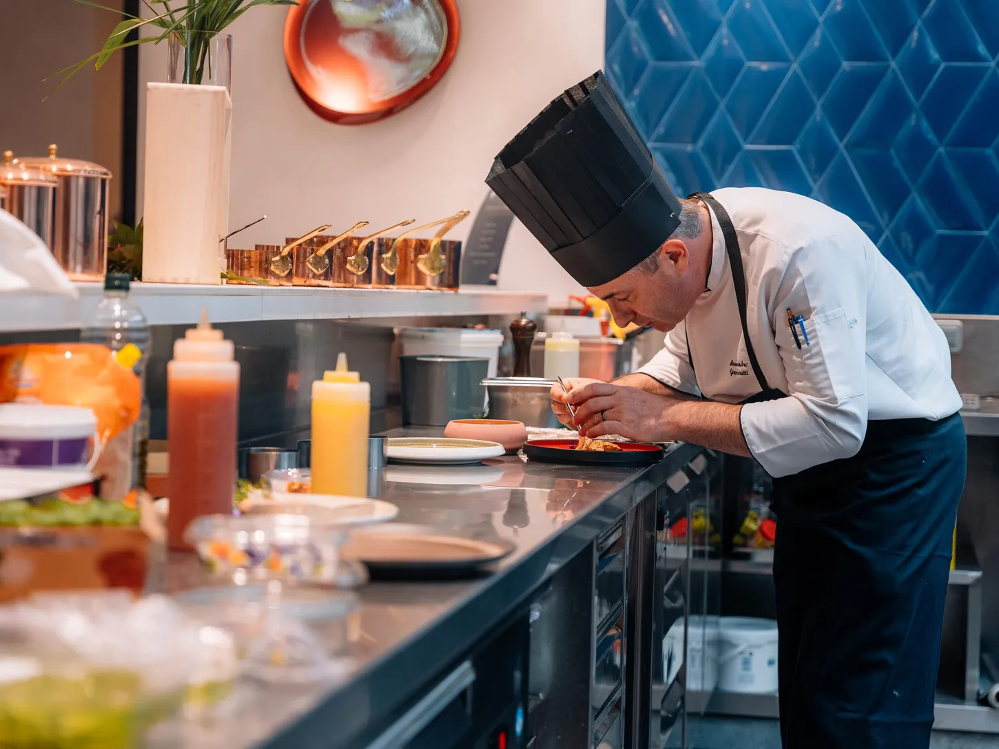 Un chef en toque blanche prépare une assiette dans une cuisine professionnelle avec divers ingrédients et ustensiles autour de lui