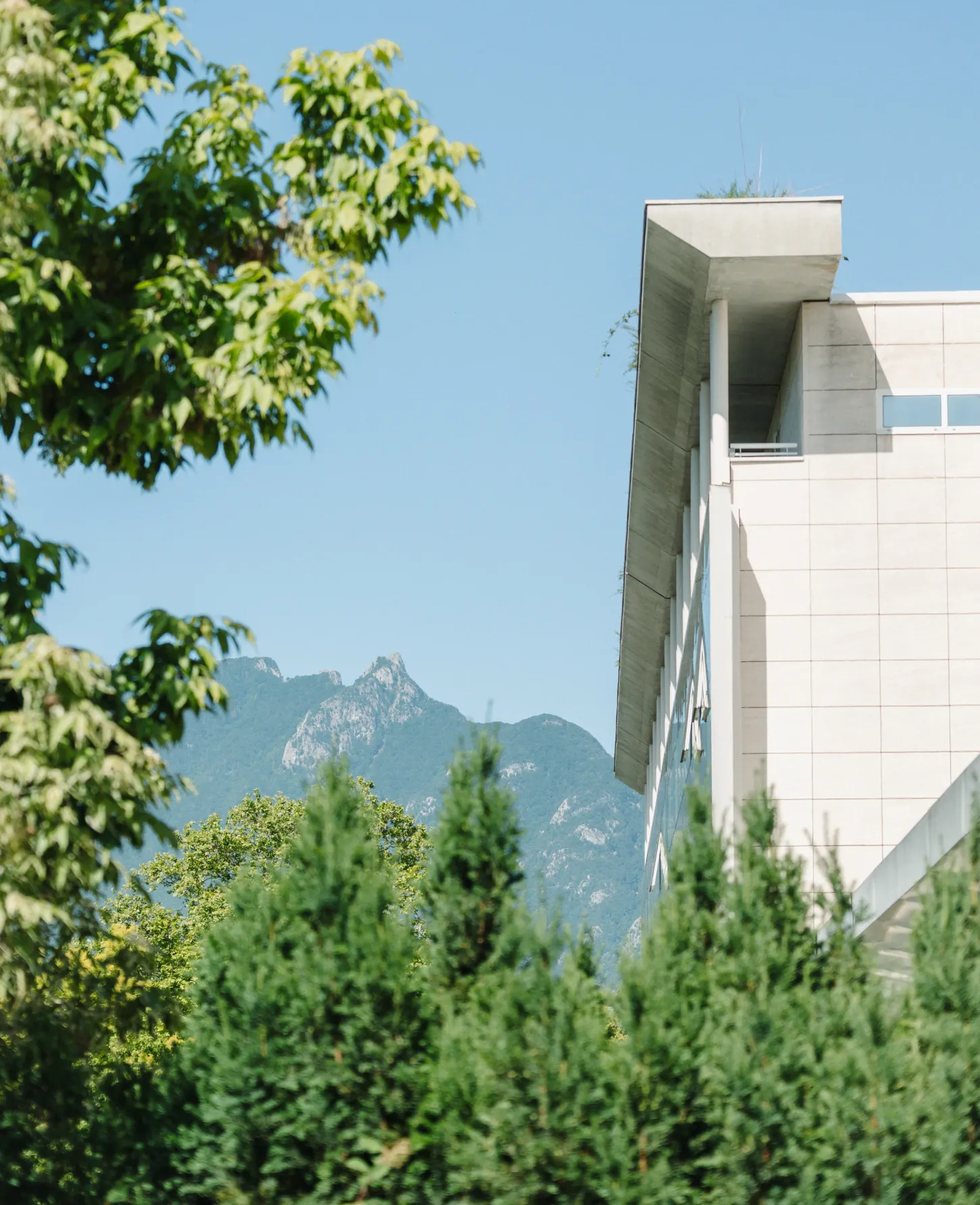 Un bâtiment moderne avec des éléments architecturaux en béton et des montagnes en arrière-plan, entouré d'arbres verts