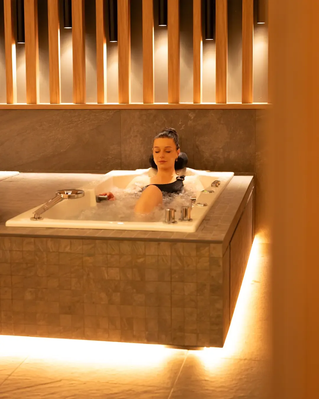 A woman is lying in a hot tub surrounded by light wood tiles and lit by golden light.
