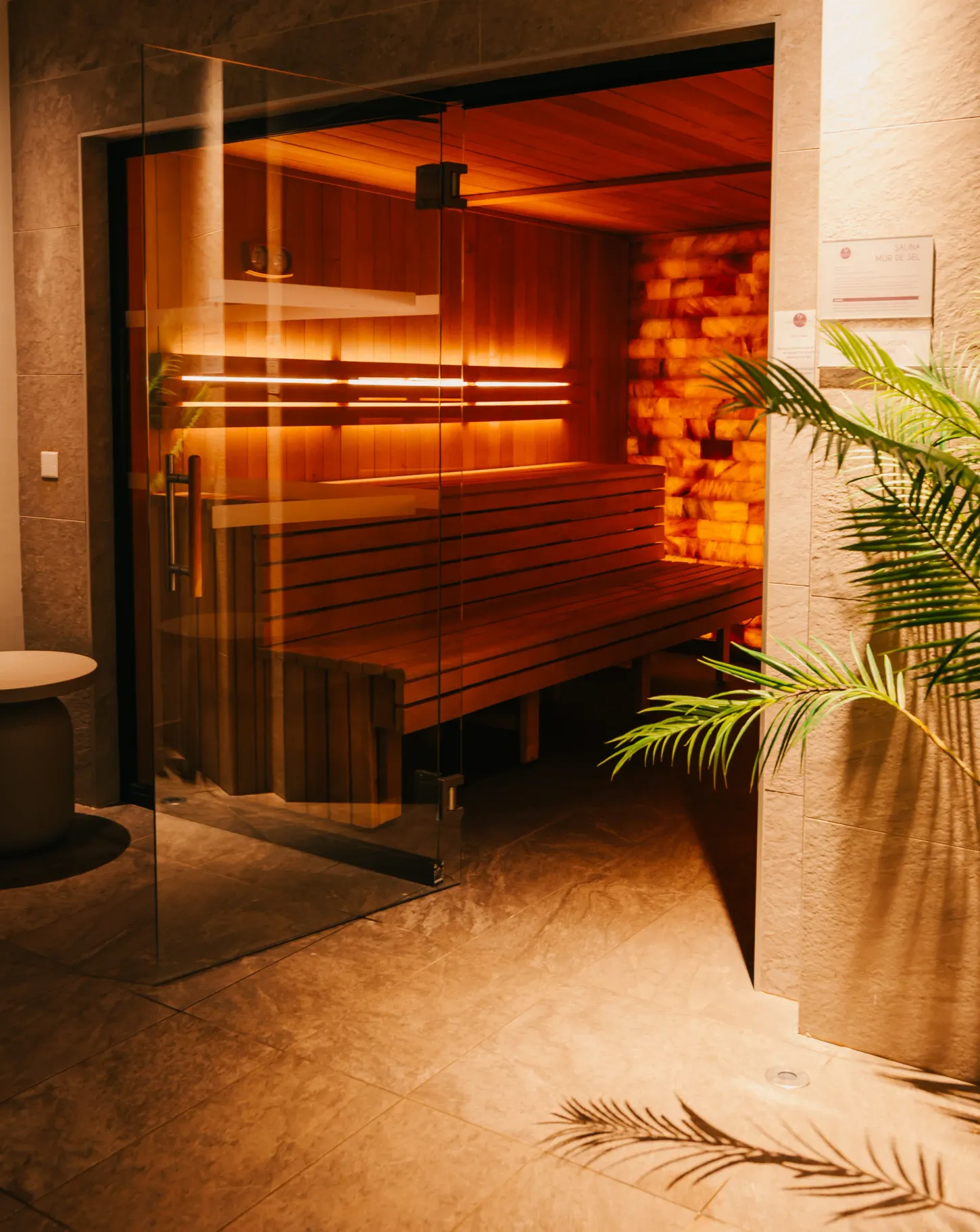 Wooden sauna room with benches, lit by warm lights, seen through an open glass door
