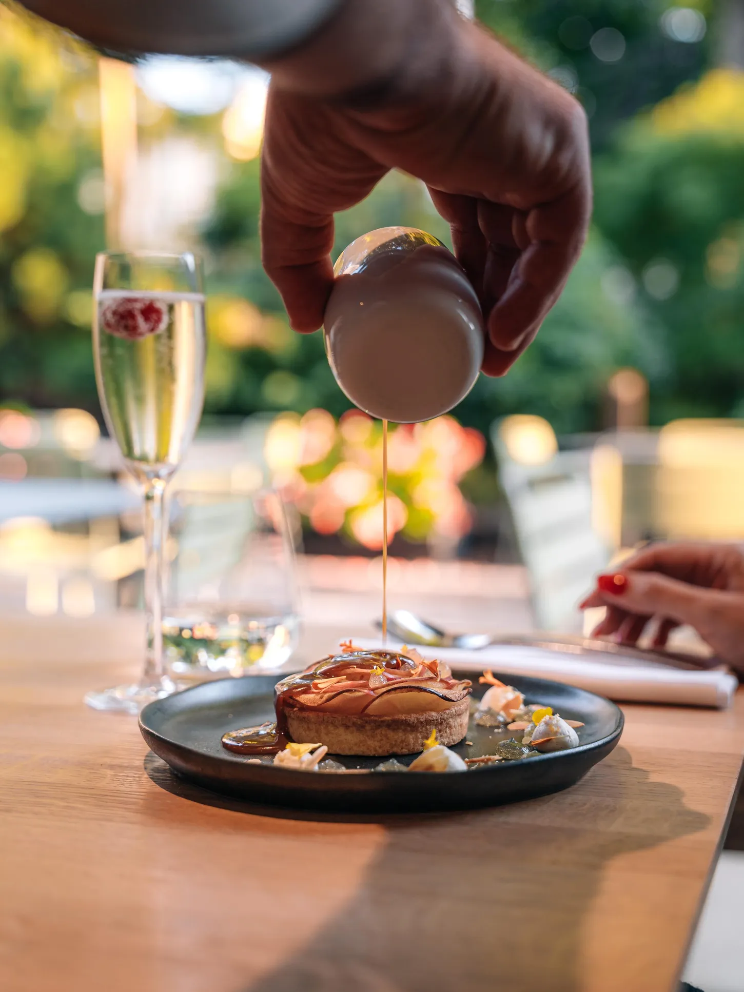 Une main verse un filet de sauce sur un plat contenant un morceau de pain garni de garnitures diverses, posé sur une table en bois avec des verres de champagne à proximité
