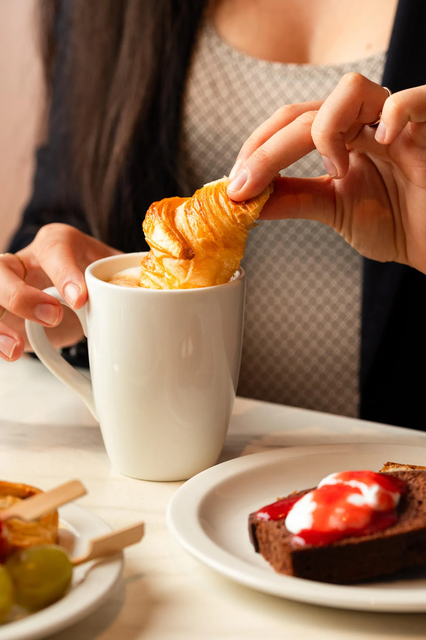 Une personne plonge un croissant dans une tasse de café, avec une assiette de tartine garnie de fromage frais et de confiture à proximité