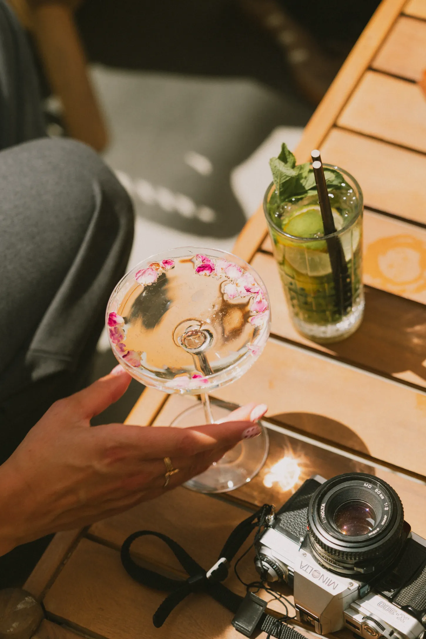 Une main tient un verre à cocktail garni de pétales de fleurs sur une table en bois, à côté d'un verre de citronnade et d'un appareil photo