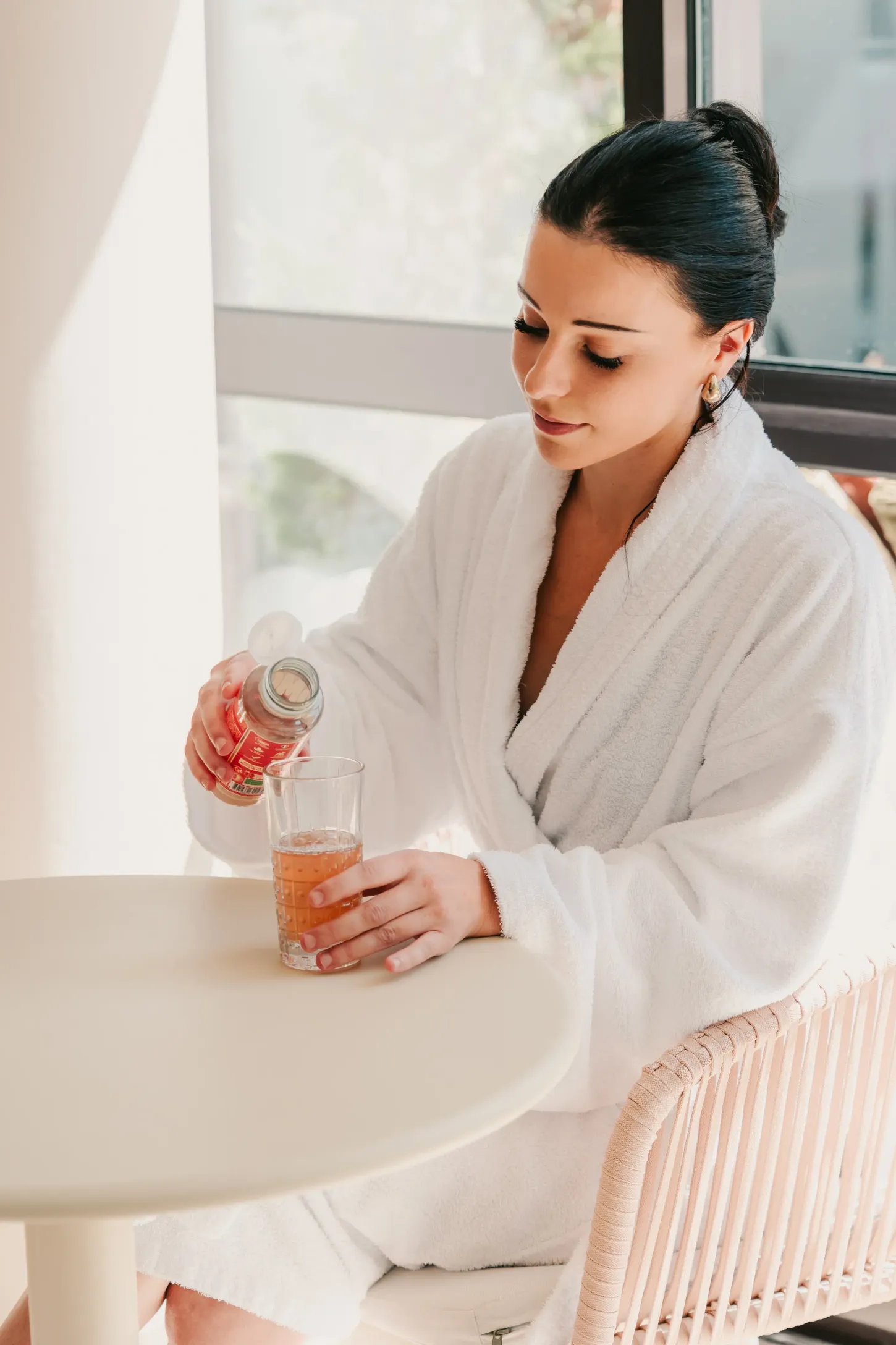 A woman in a white bathing robe pours a liquid into a glass on a table near a window.