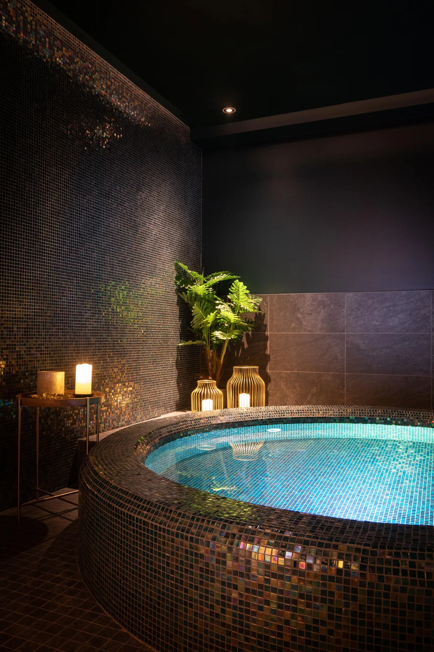 Jacuzzi intérieur avec éclairage bleu, entouré de mosaïques et de plantes, avec des bougies et une lampe à proximité
