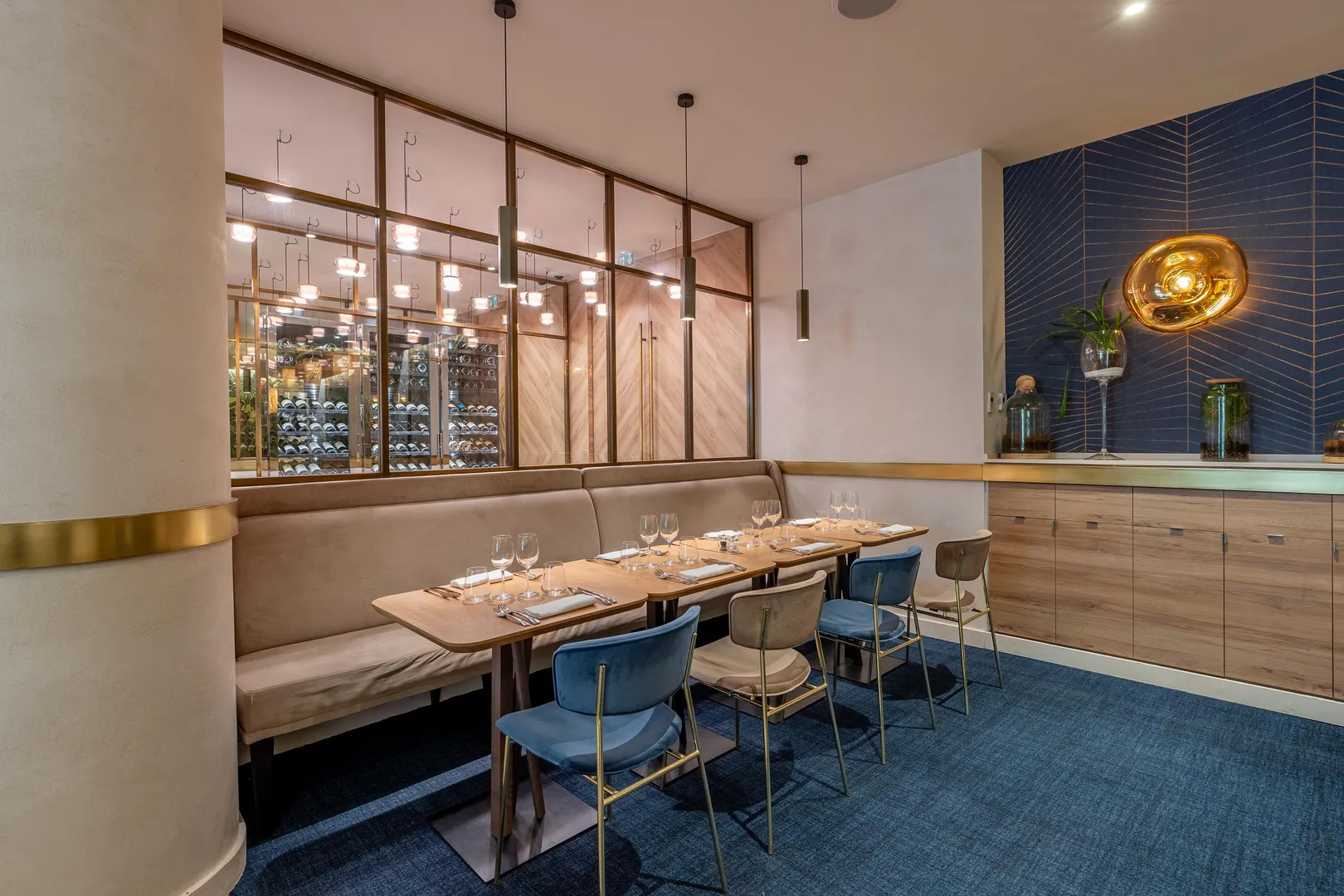 Un restaurant élégant avec une table dressée, des chaises bleues et une banquette, devant une vitrine à vin éclairée