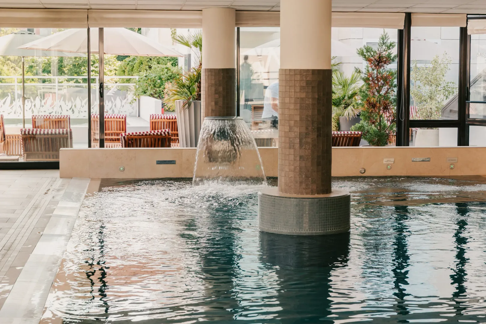 Indoor pool with a central fountain and tables and chairs on a terrace visible through the large glass windows.