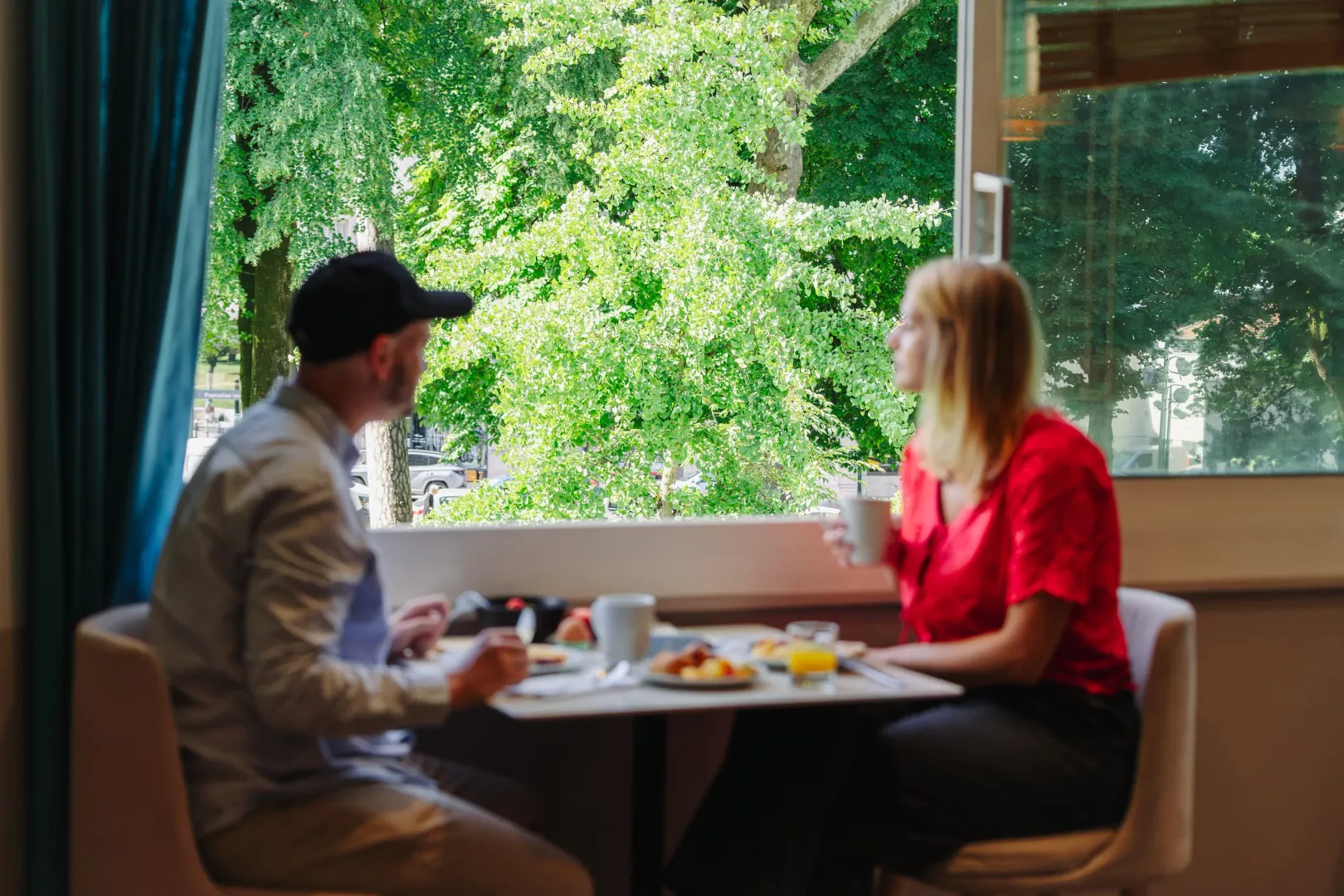 Two people sitting at a table with food and drinks, talking in an indoor setting with a large window overlooking flowering trees.