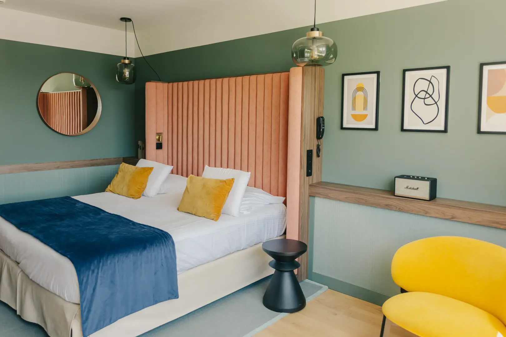 Zimmer mit einem weißen und blauen Bett, gelben Kissen, einem orangen Bettkopf, einer Wandlampe, Bildern an den Wänden und einem gelben Sessel