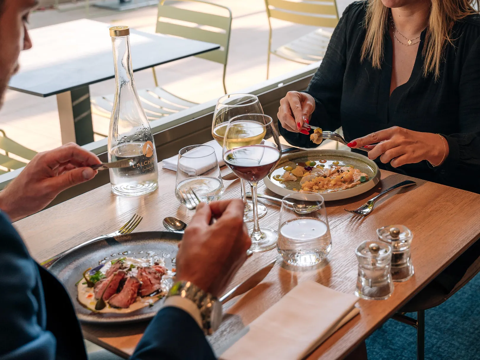 Trois personnes assises à une table en train de manger des plats et de boire du vin, avec des verres, des couverts et des bouteilles sur la table