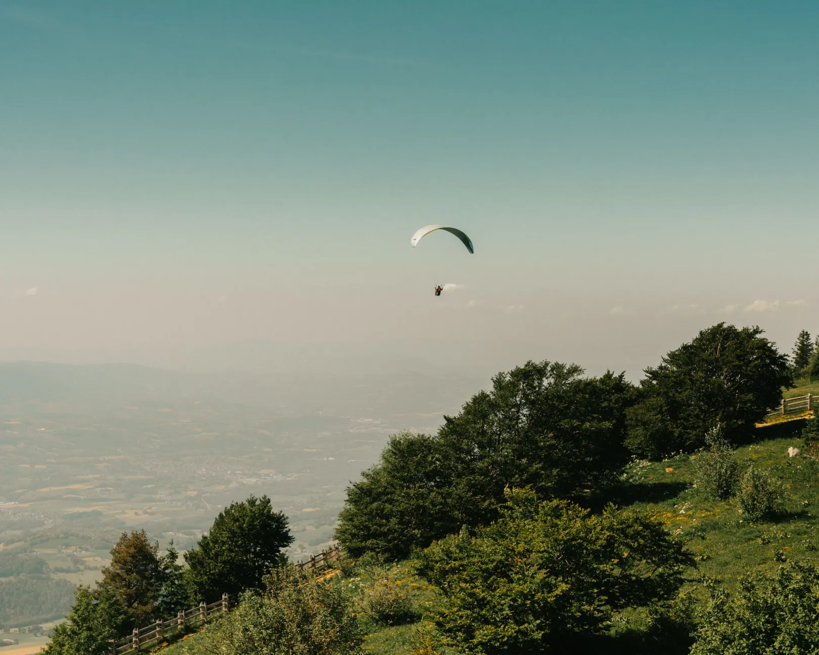 A paraglider flies above a wooded hill with a view of a valley below.