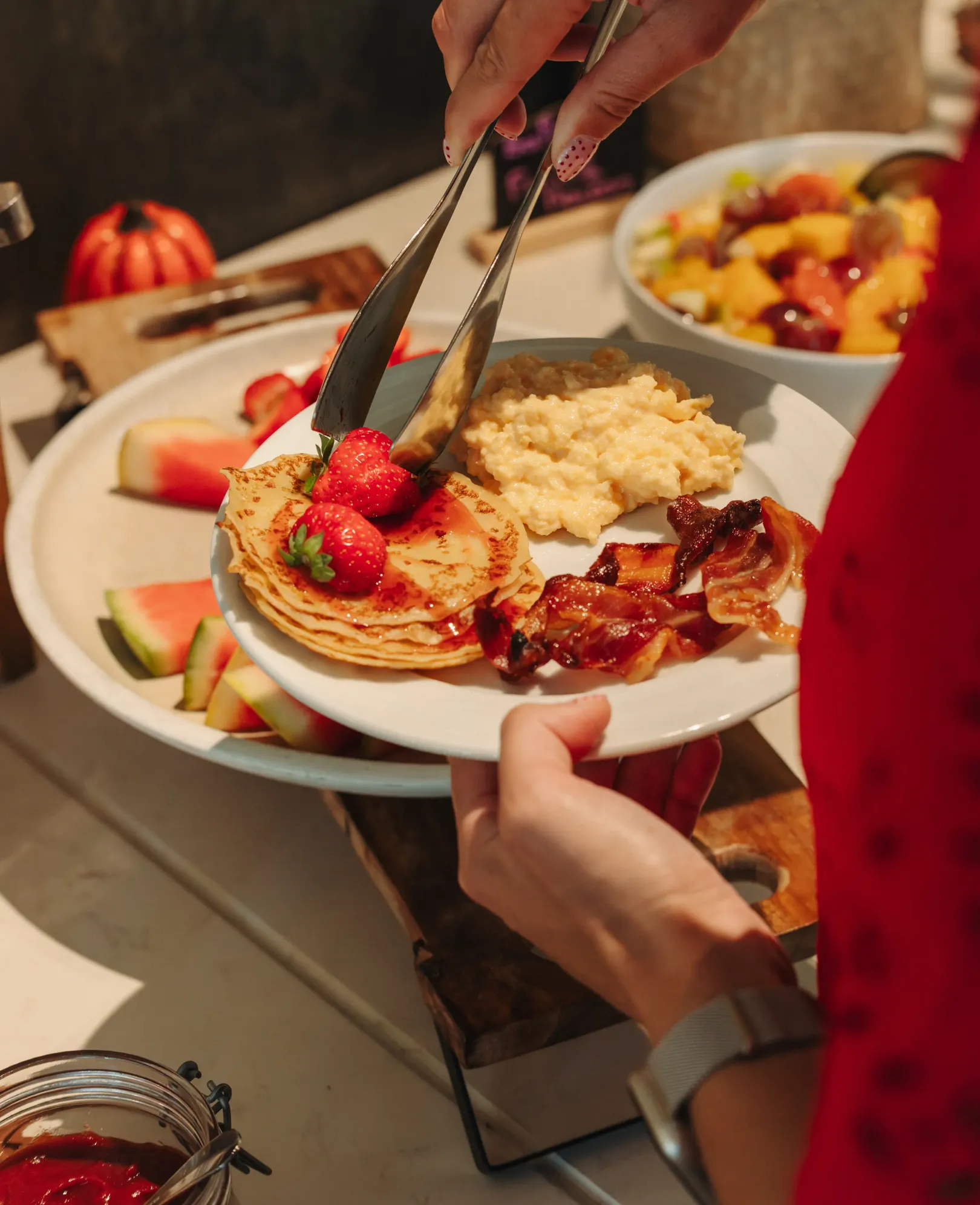 A person serves pancakes topped with strawberries, bacon, and fruit puree on a plate, surrounded by fruit and other dishes.
