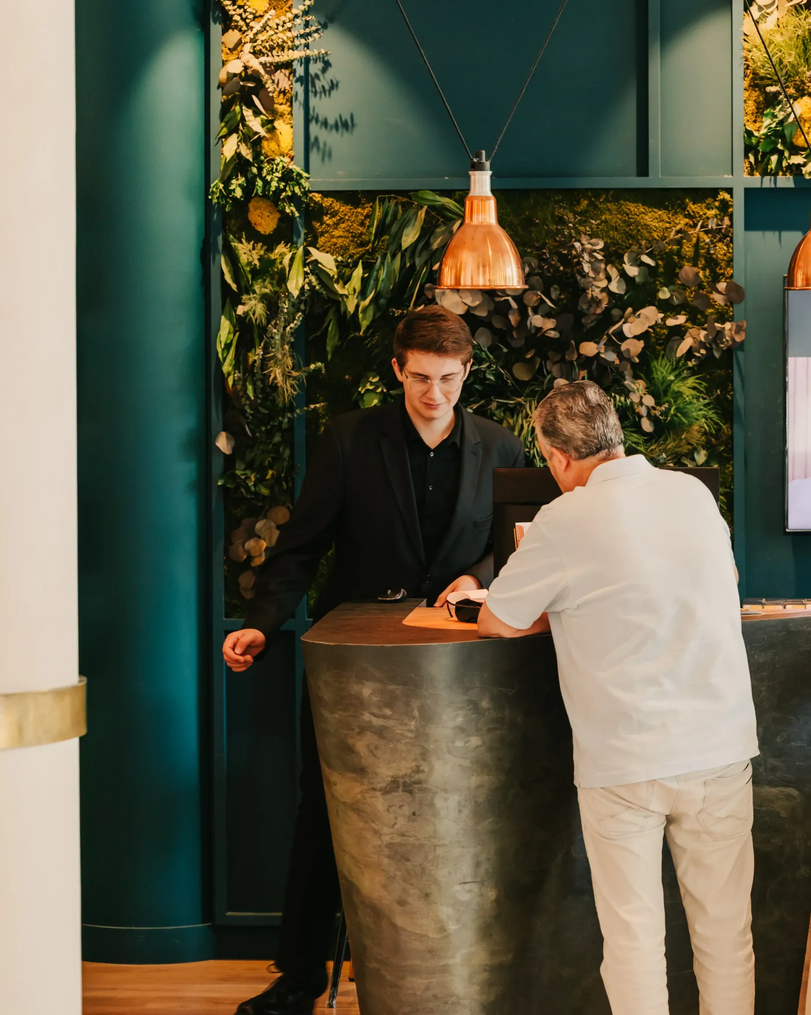 Two men are talking at a counter in front of a wall decorated with green plants and flowers.