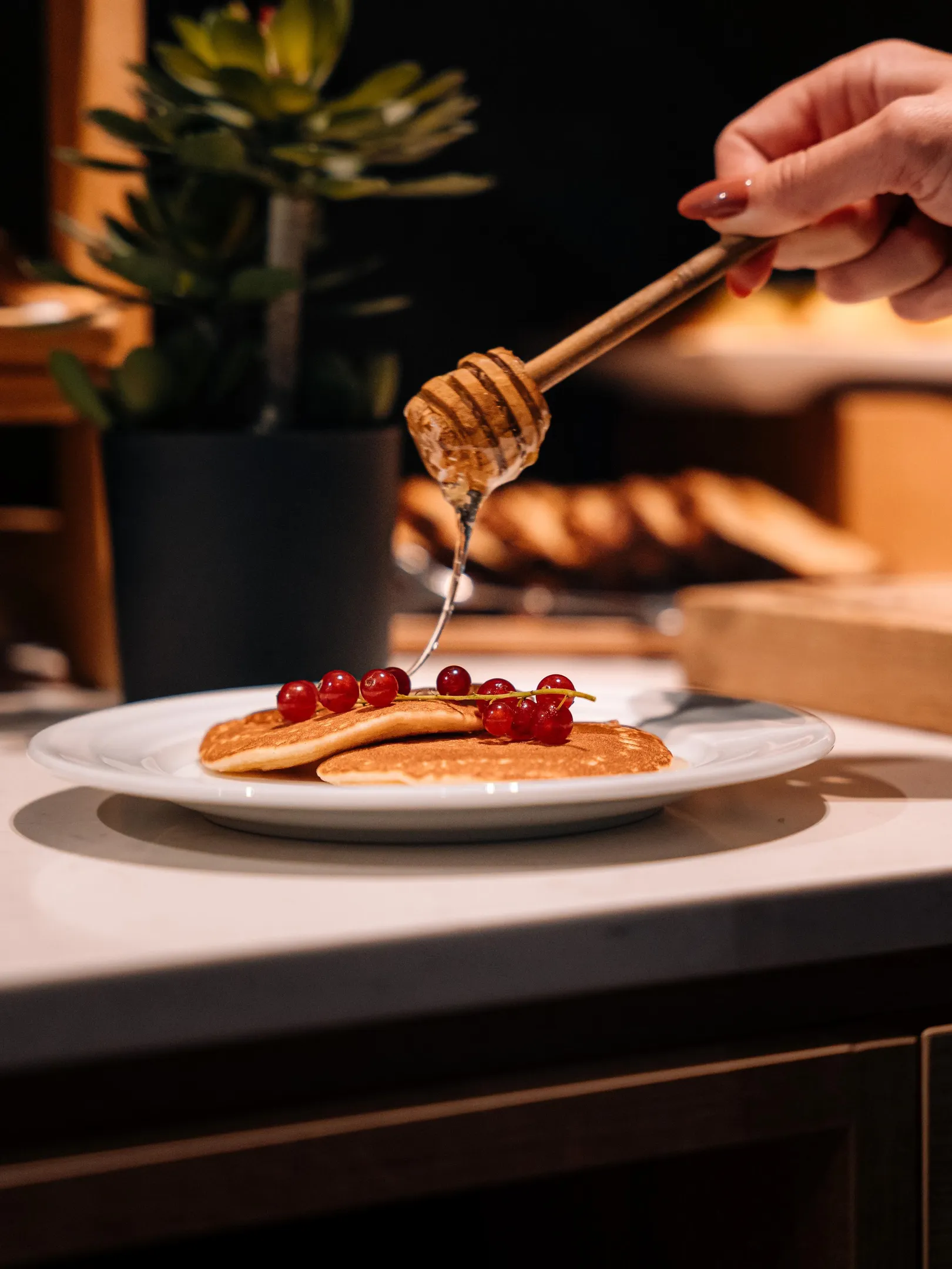 Une main verse du miel sur des pancakes garnis de baies rouges posés sur une assiette blanche