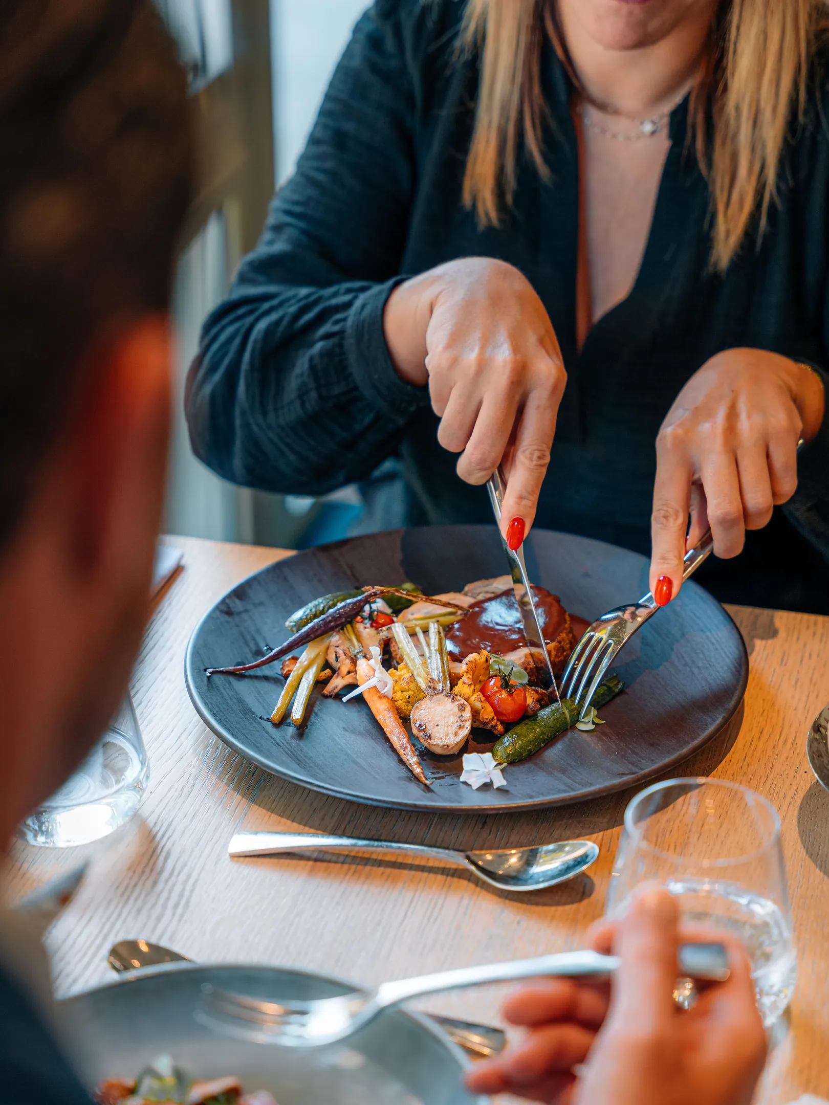 Une personne coupe un plat avec des légumes et une sauce à l'aide d'une fourchette et d'un couteau sur une assiette noire posée sur une table en bois