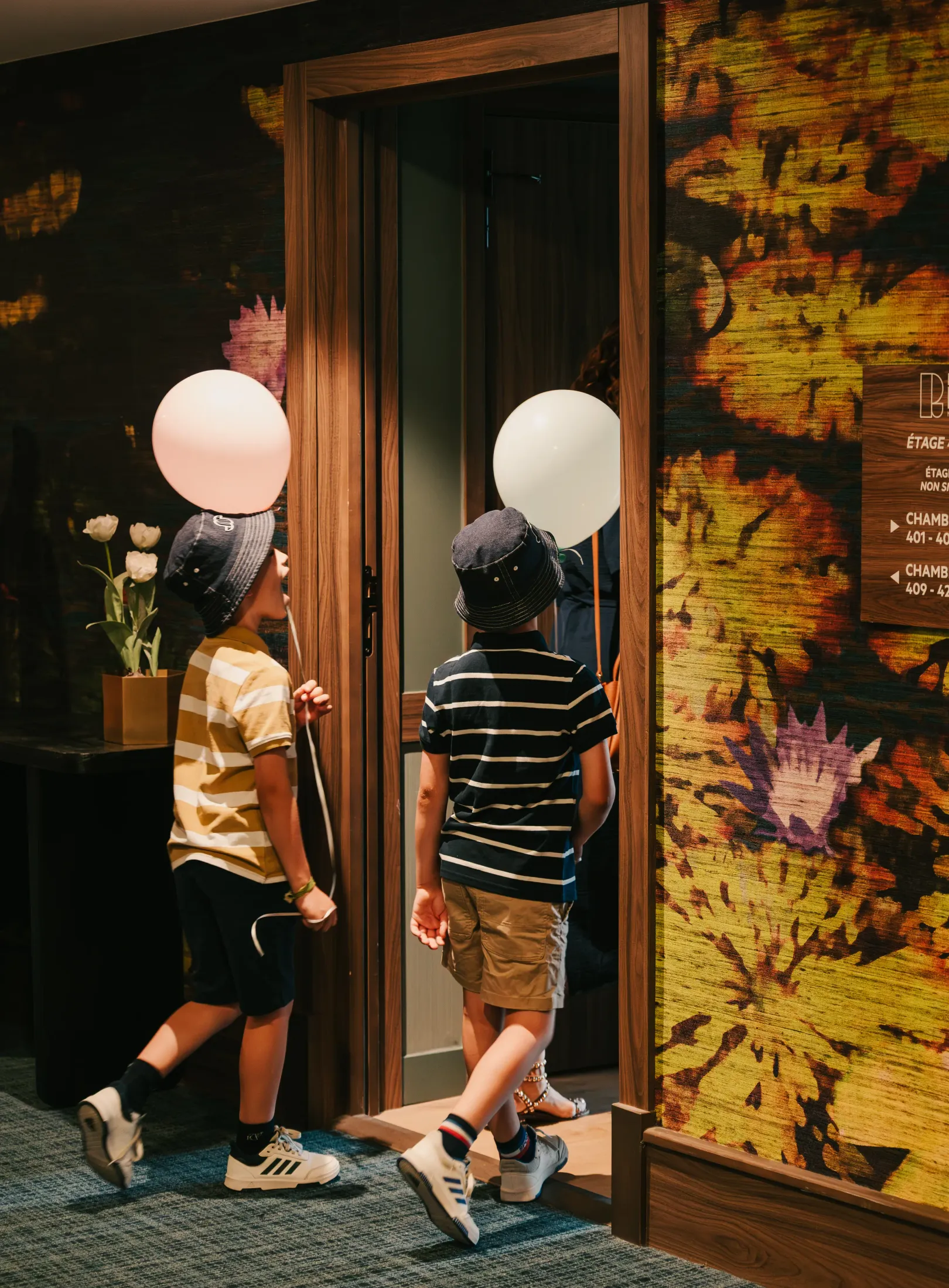Two children wearing hats and white balloons walk in front of a wooden door with a wall decorated with colorful patterns next to it.