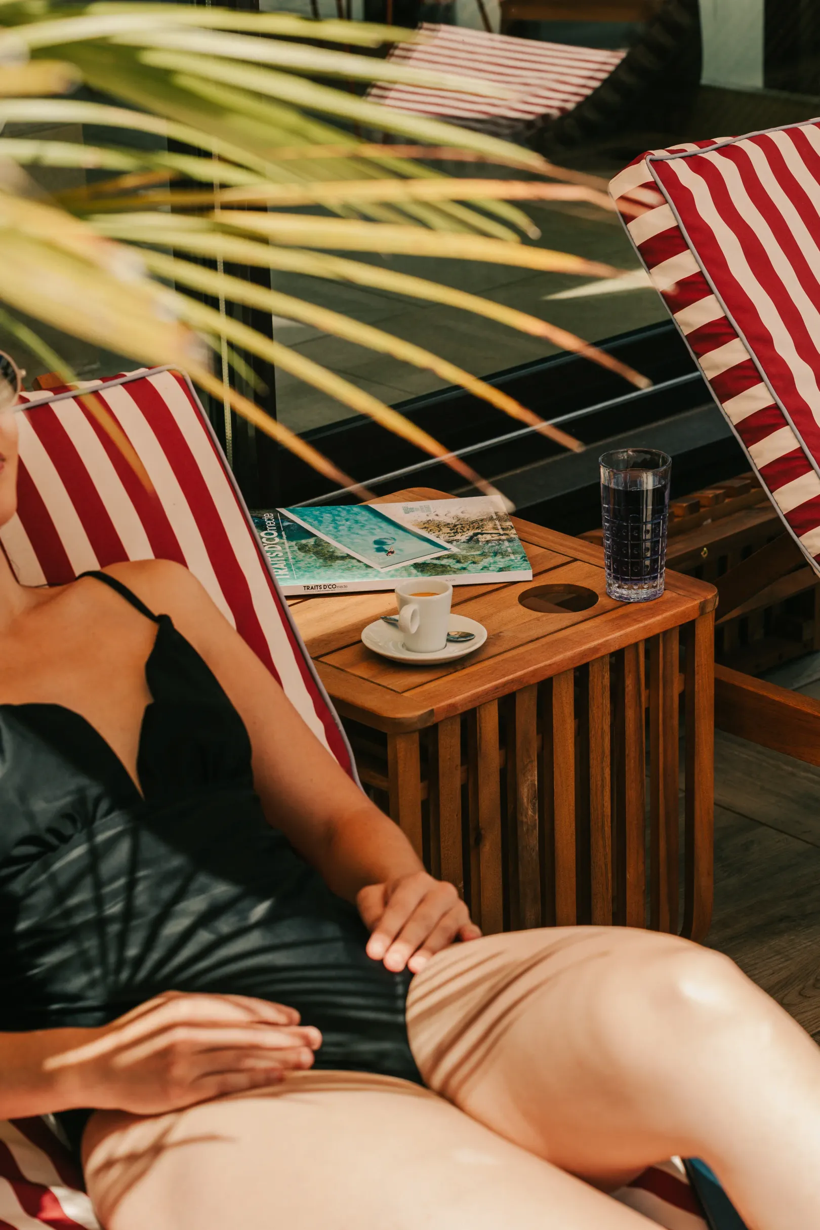 Two people lying on sun loungers hold hands, with a table containing a coffee and magazines between them.