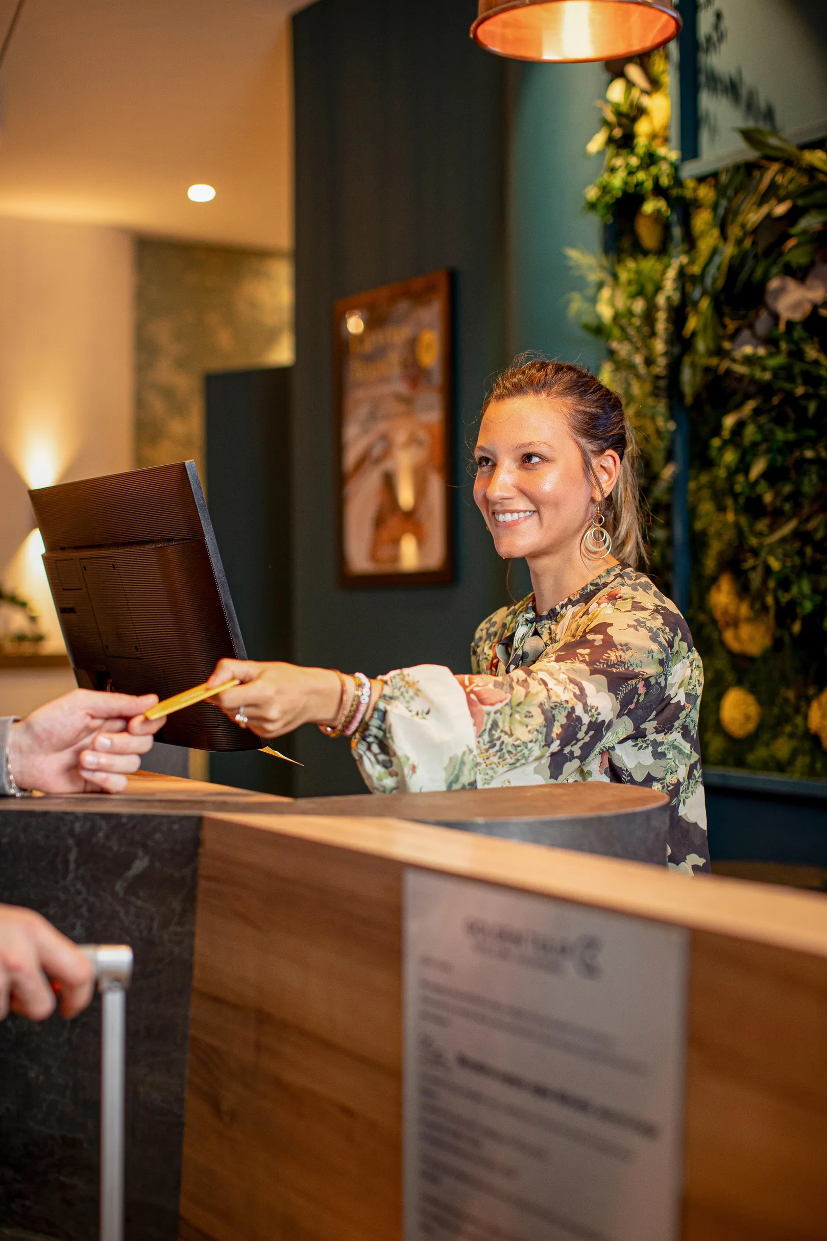 Une femme derrière un comptoir remet une carte à un client dans un hall d'hôtel décoré de plantes