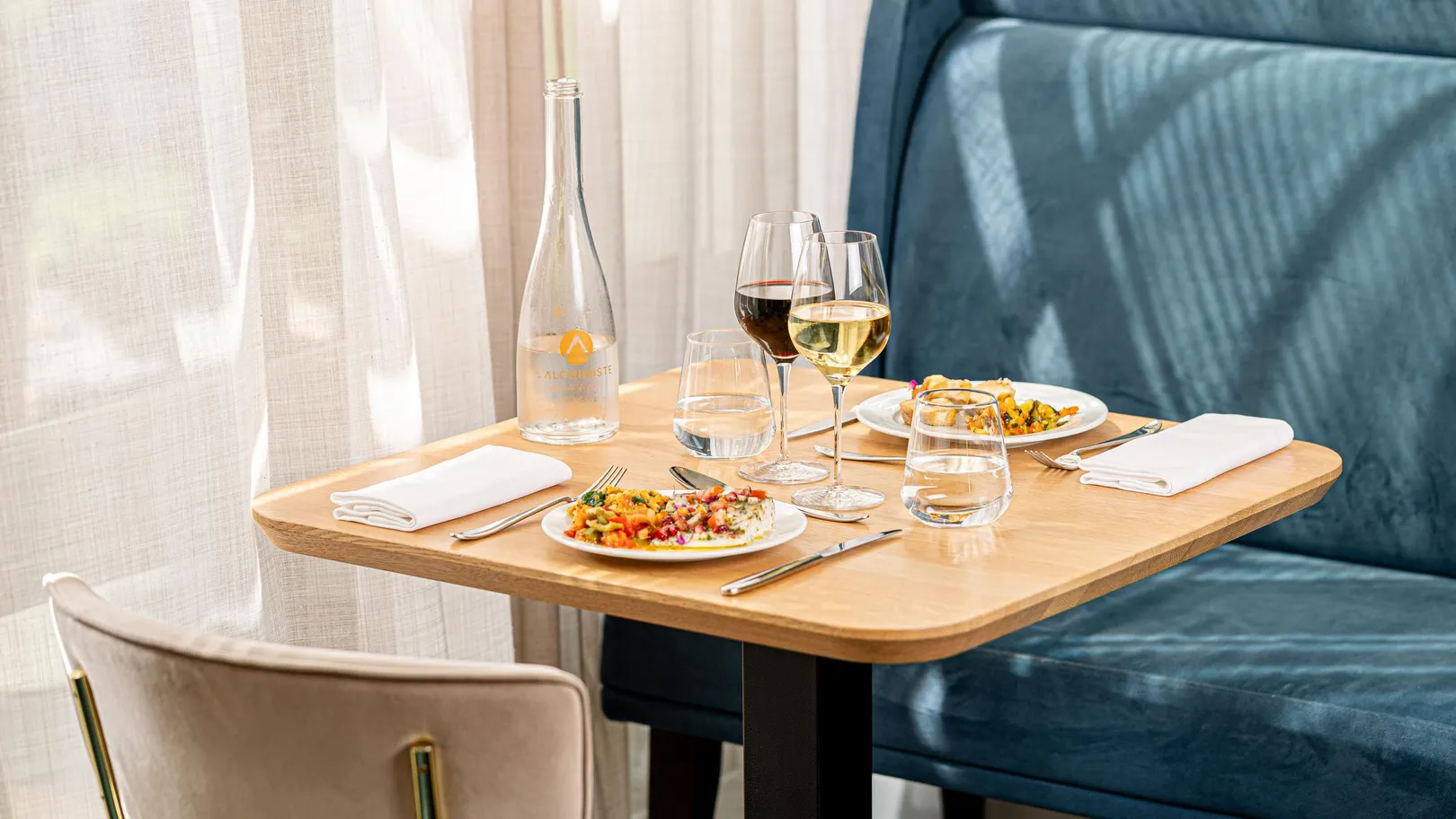 Tisch gedeckt mit zwei Tellern Essen, zwei Weingläsern, einer Wasserflasche und Wassergläsern, nahe einem Fenster mit weißen Vorhängen