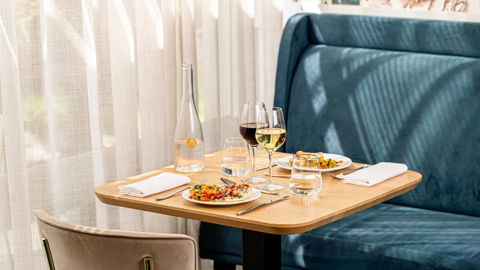 Table set with two salad plates, two wine glasses, two water glasses, one water bottle, and a white tablecloth against a backdrop of a blue bench and white curtains.