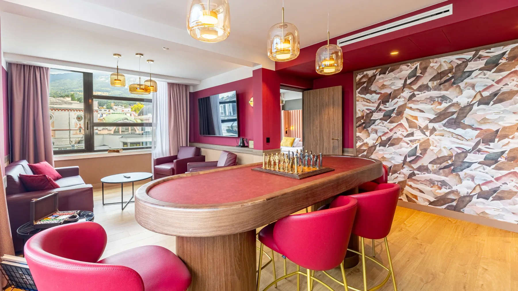Salle de séjour avec table ovale en bois, chaises rouges et canapés, décorée de luminaires et d'un mur mural représentant des montagnes
