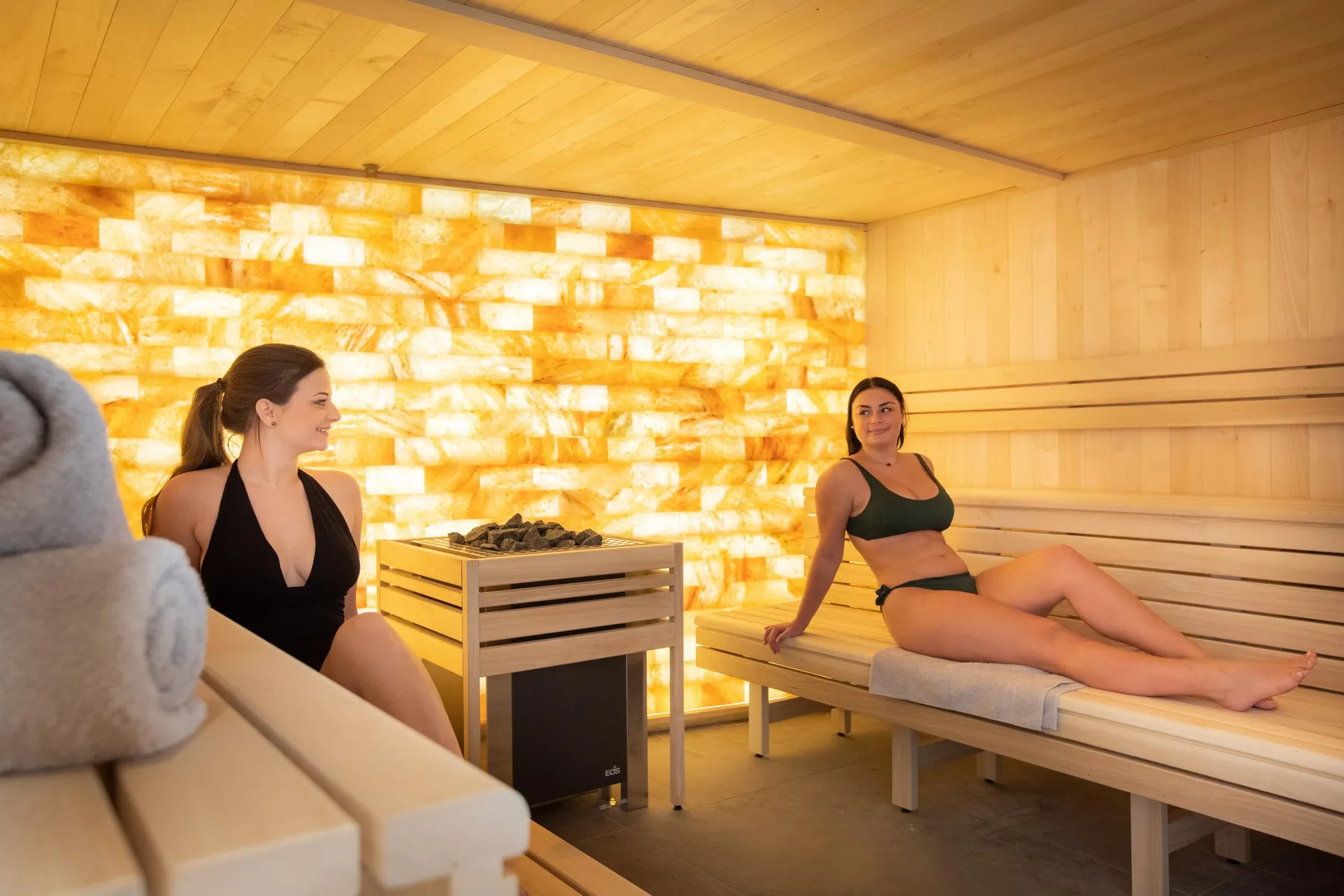 Deux femmes assises dans un sauna en bois éclairé par une paroi lumineuse, l'une en maillot noir et l'autre en tenue verte
