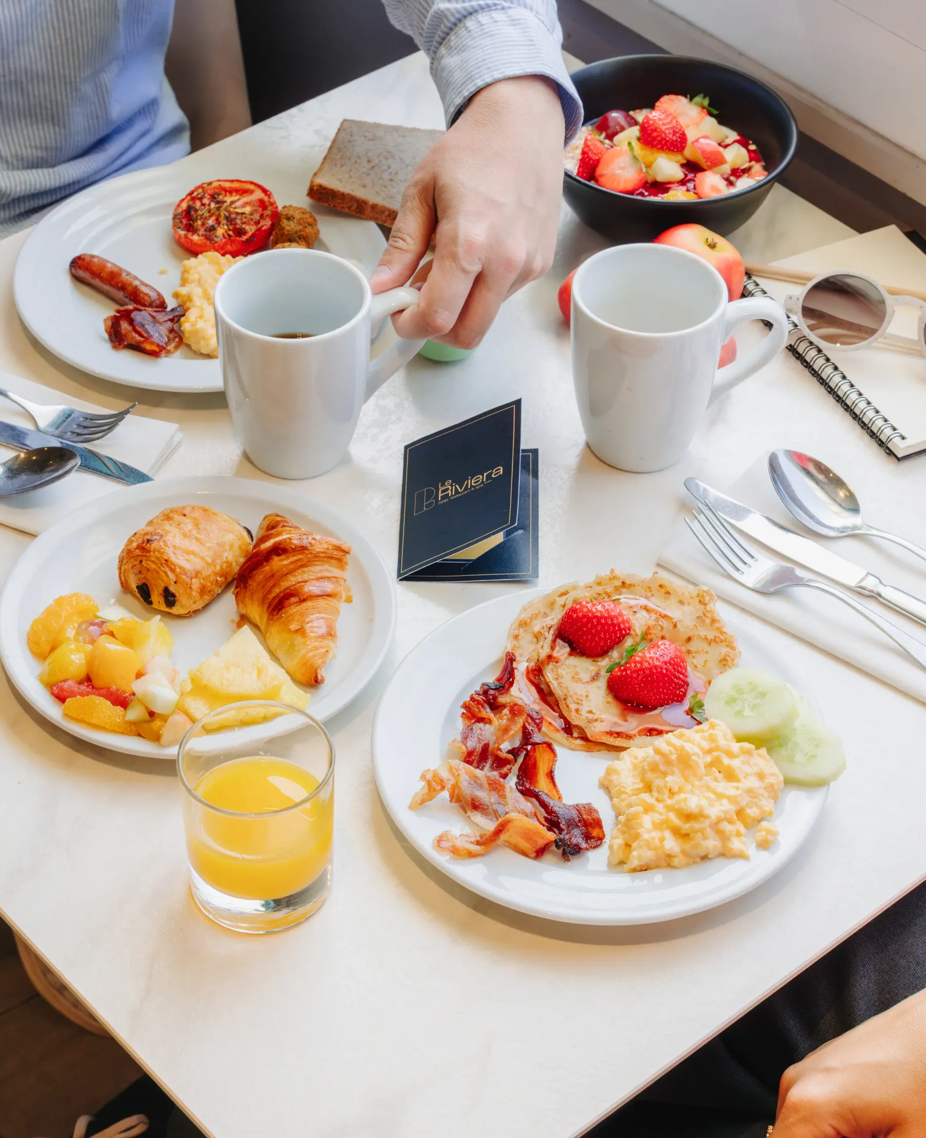 Eine Person nimmt ein Croissant von einem Tisch mit verschiedenen Frühstücksgerichten, Getränken und Früchten.