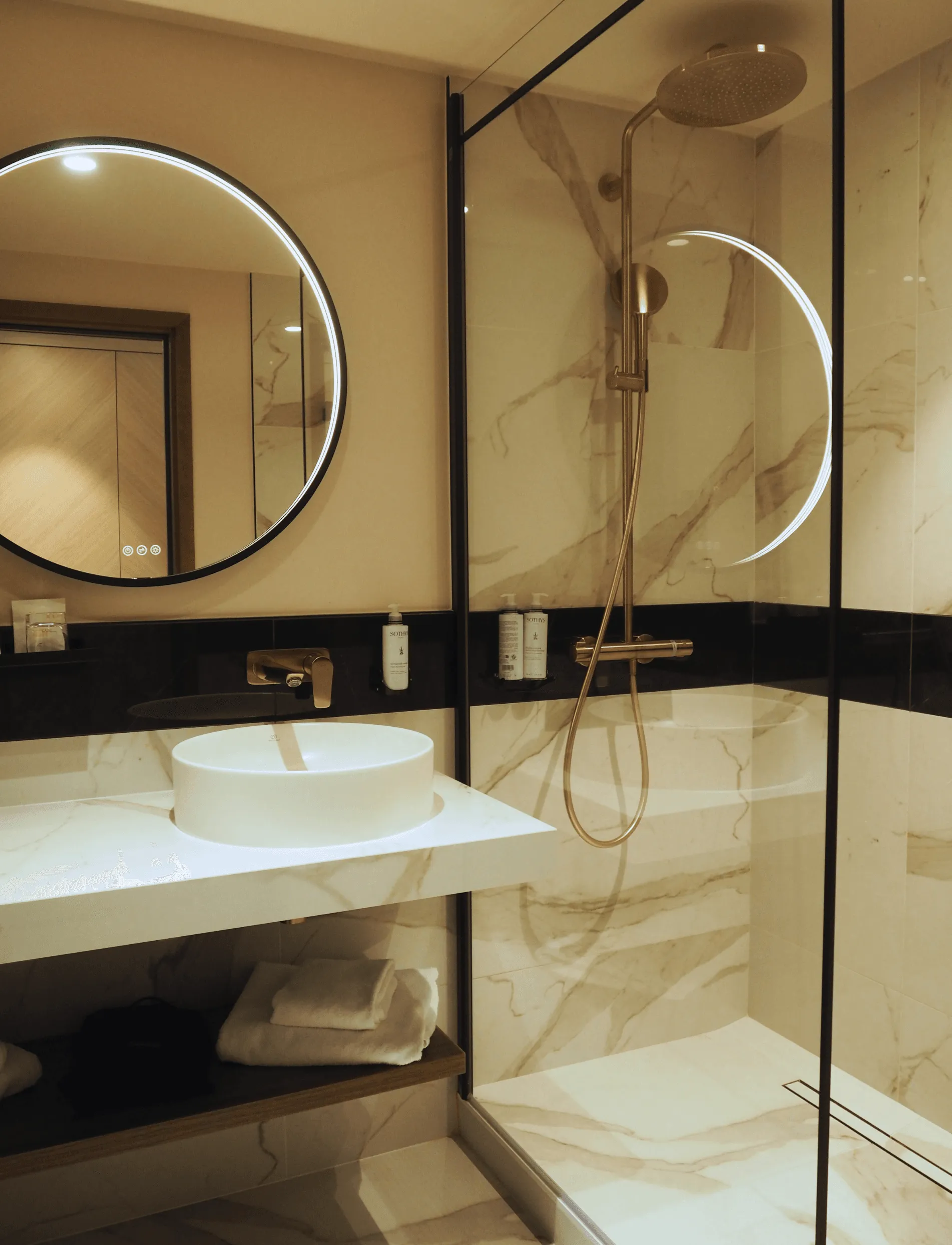 Modernes Badezimmer mit weißem Waschbecken, beleuchtetem Rundspiegel und Regendusche hinter einer Glasabtrennung