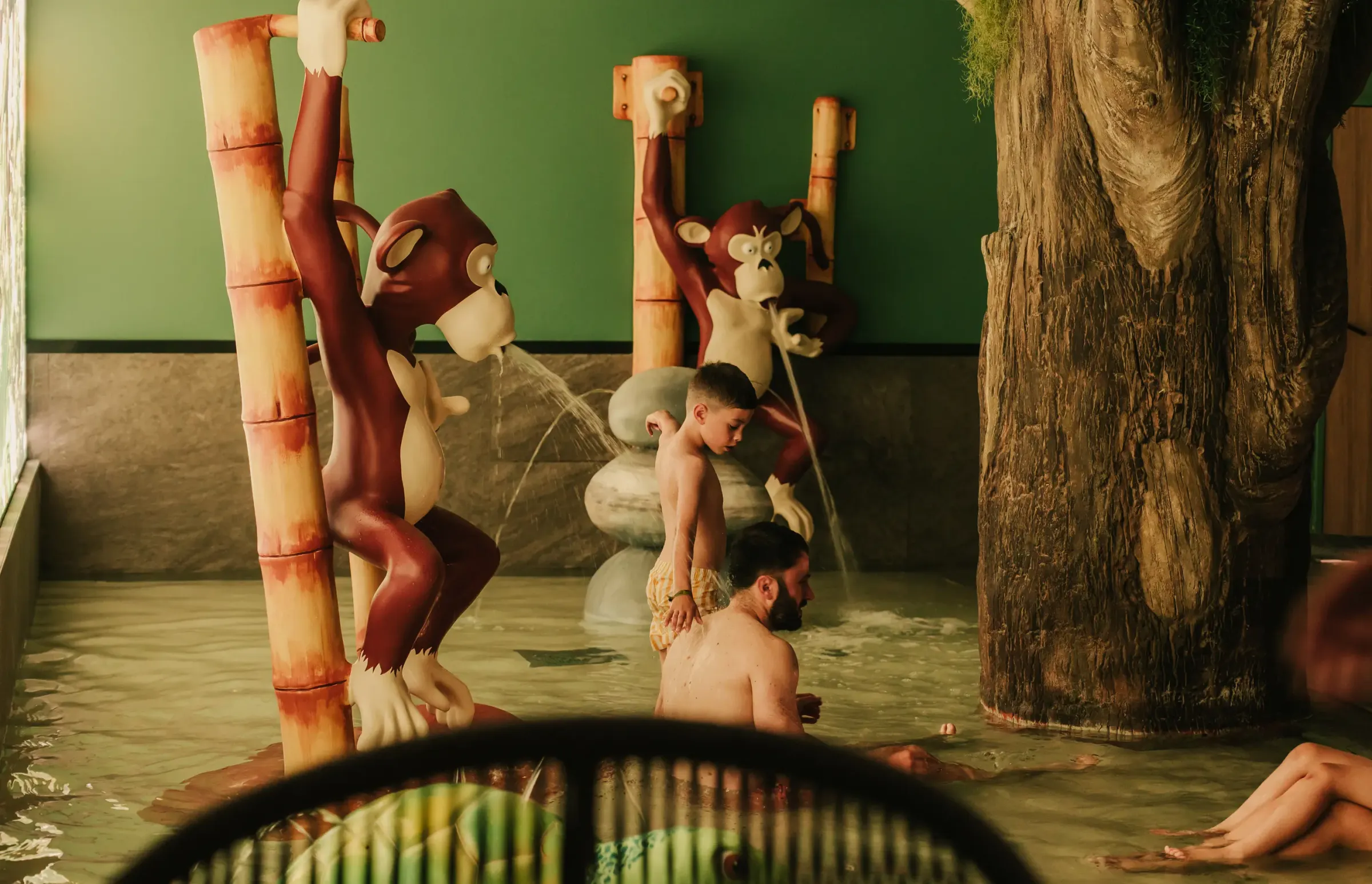 People relax in a thermal bath decorated with monkey sculptures and bamboo, with water jets in the background.