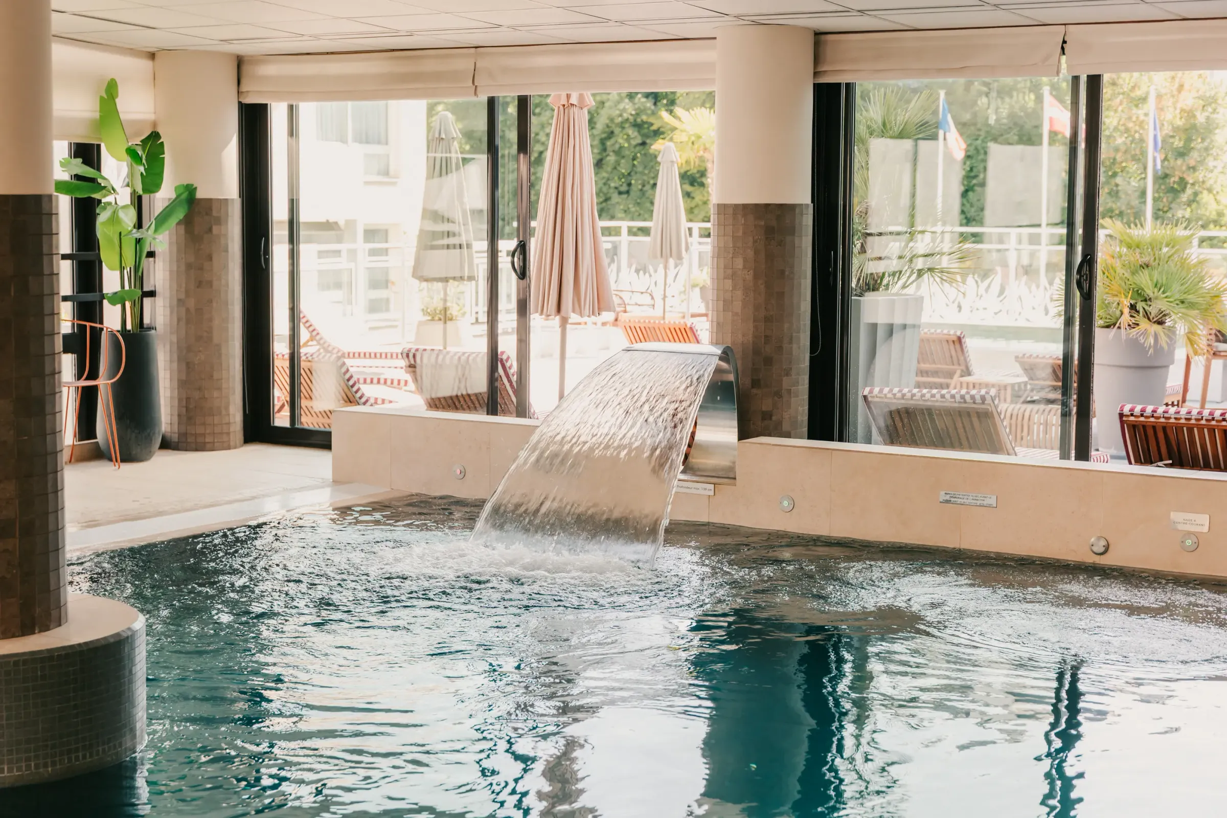 Indoor pool with a small basin and a waterfall, surrounded by large glass windows opening onto a terrace with sun loungers