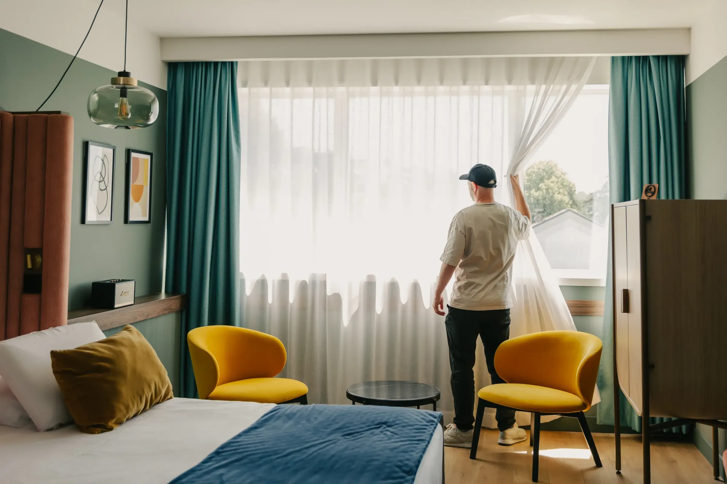 Un homme ouvre les rideaux d'une grande fenêtre dans une chambre moderne avec deux chaises jaunes et un lit