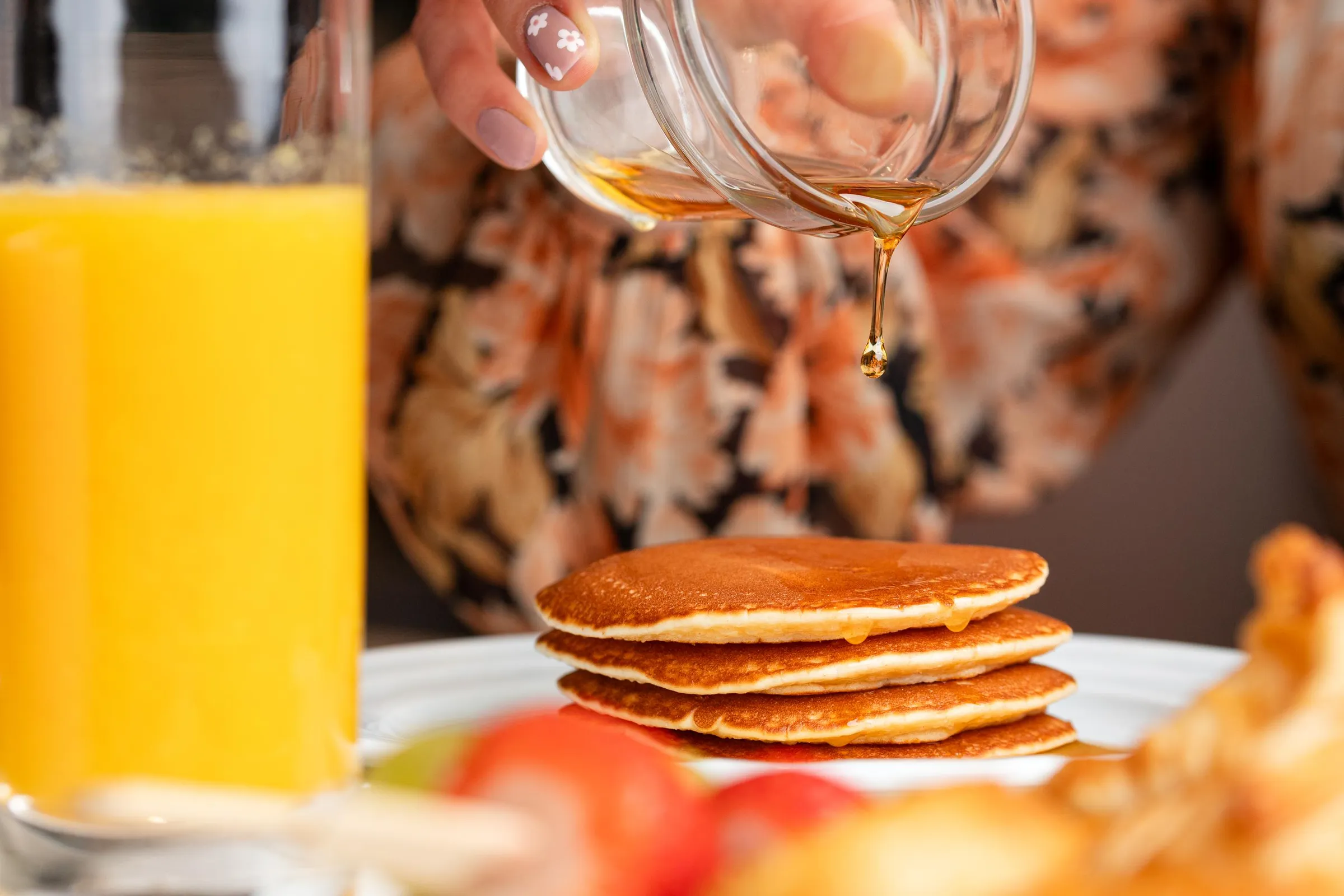 Une main verse du sirop sur une pile de pancakes, avec un verre de jus d'orange à gauche et des fruits en arrière-plan