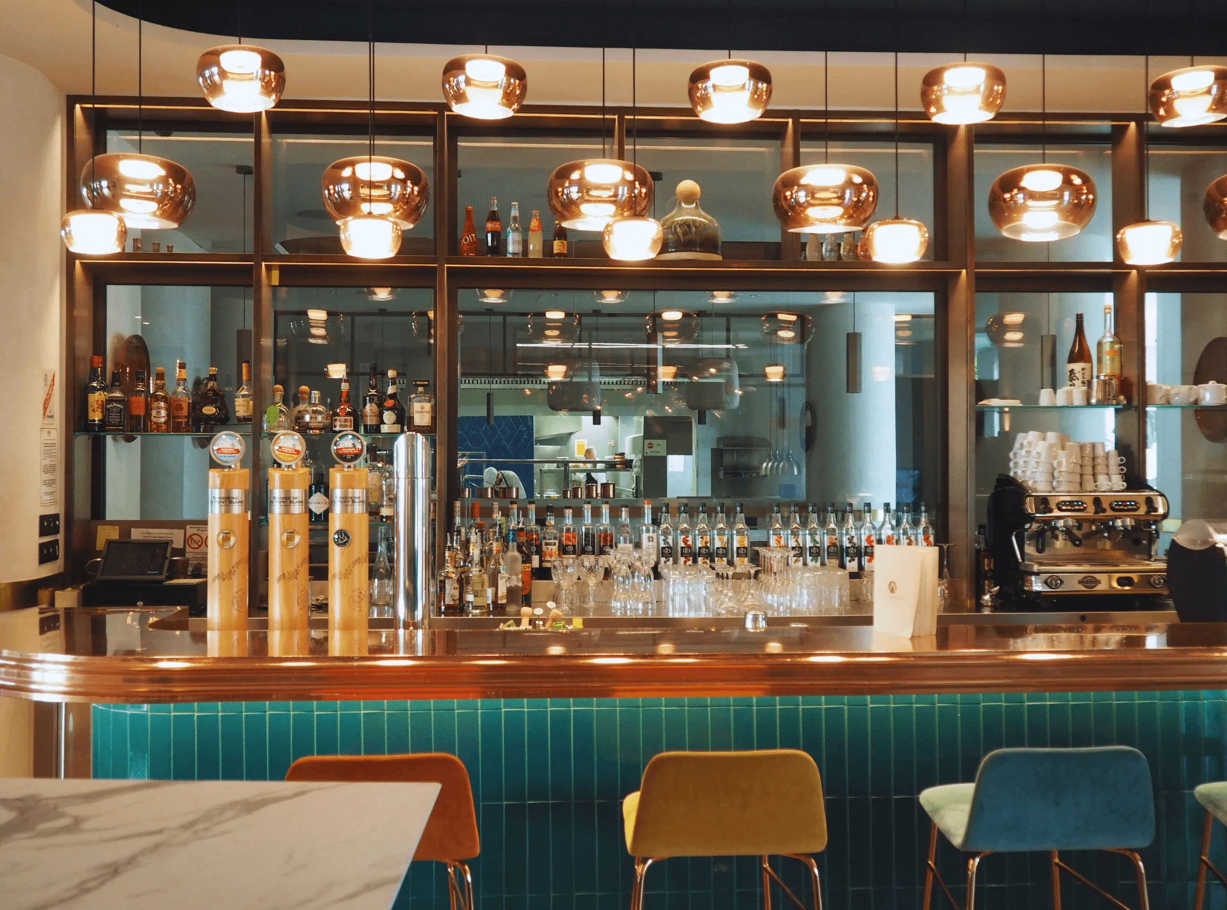 Bar avec étagères remplies de bouteilles d'alcool, luminaires en métal suspendus, et comptoir avec tabourets devant un mur carrelé vert