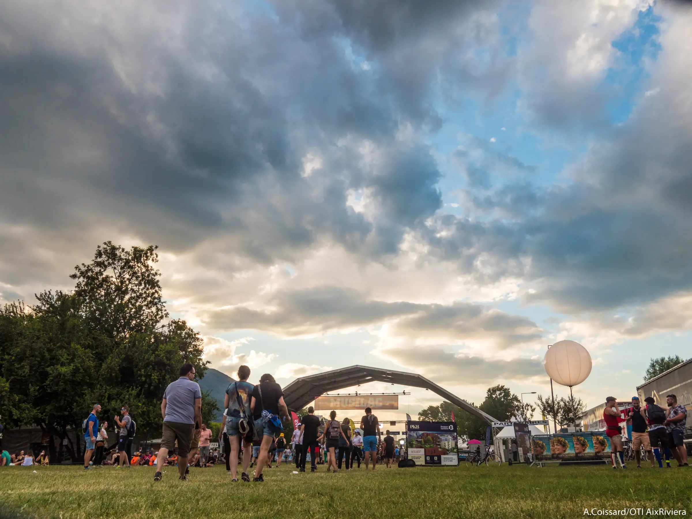 A group of people stands on a lawn in front of a concert stage under a cloudy sky with a white pear-shaped balloon nearby.