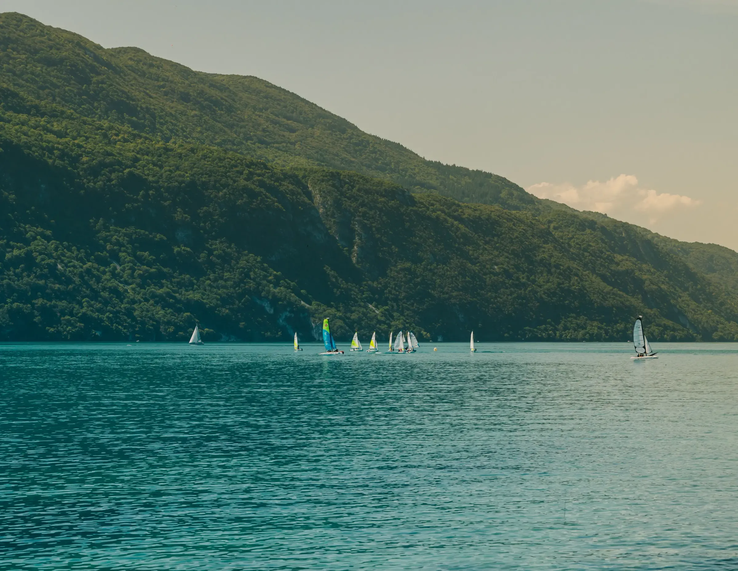 Sailboats navigate on a lake surrounded by wooded hills under a clear sky.