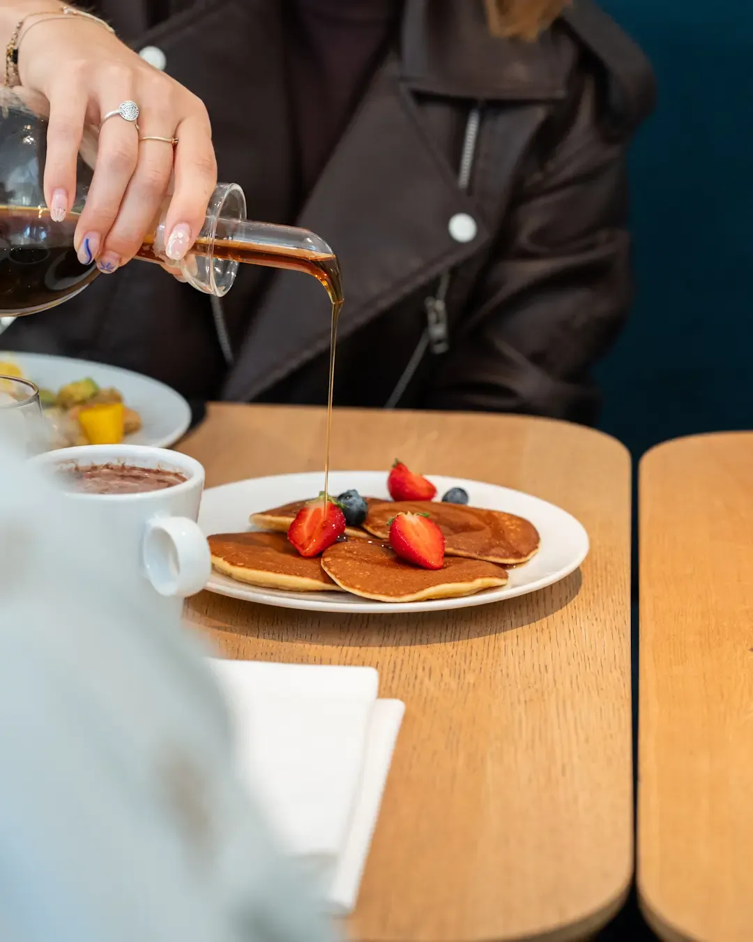 A person pours syrup over pancakes topped with red berries, accompanied by a hot drink on a wooden table.
