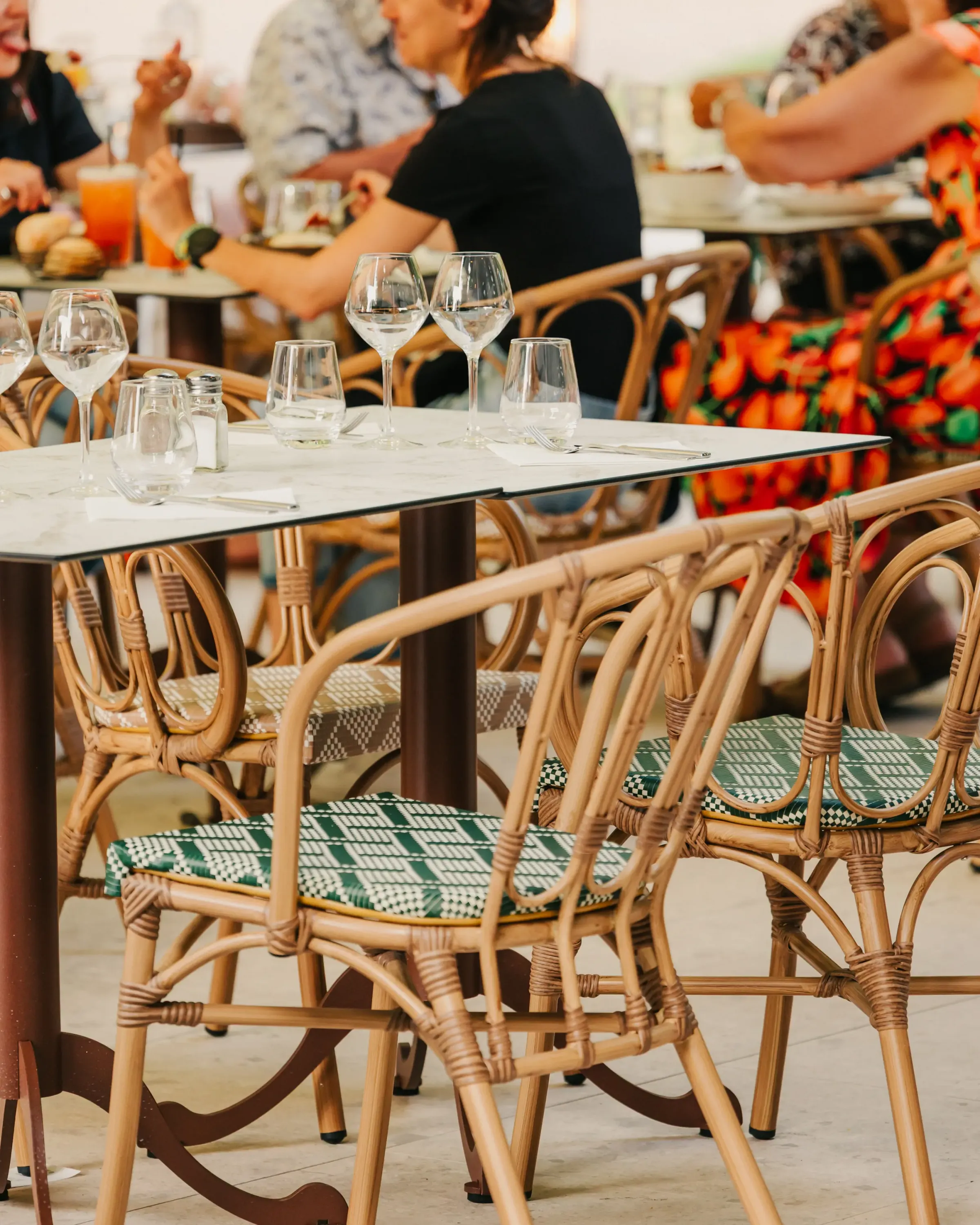 Une table en verre avec des verres et des sets de table dans un restaurant bondé avec des chaises en rotin et des clients en arrière-plan