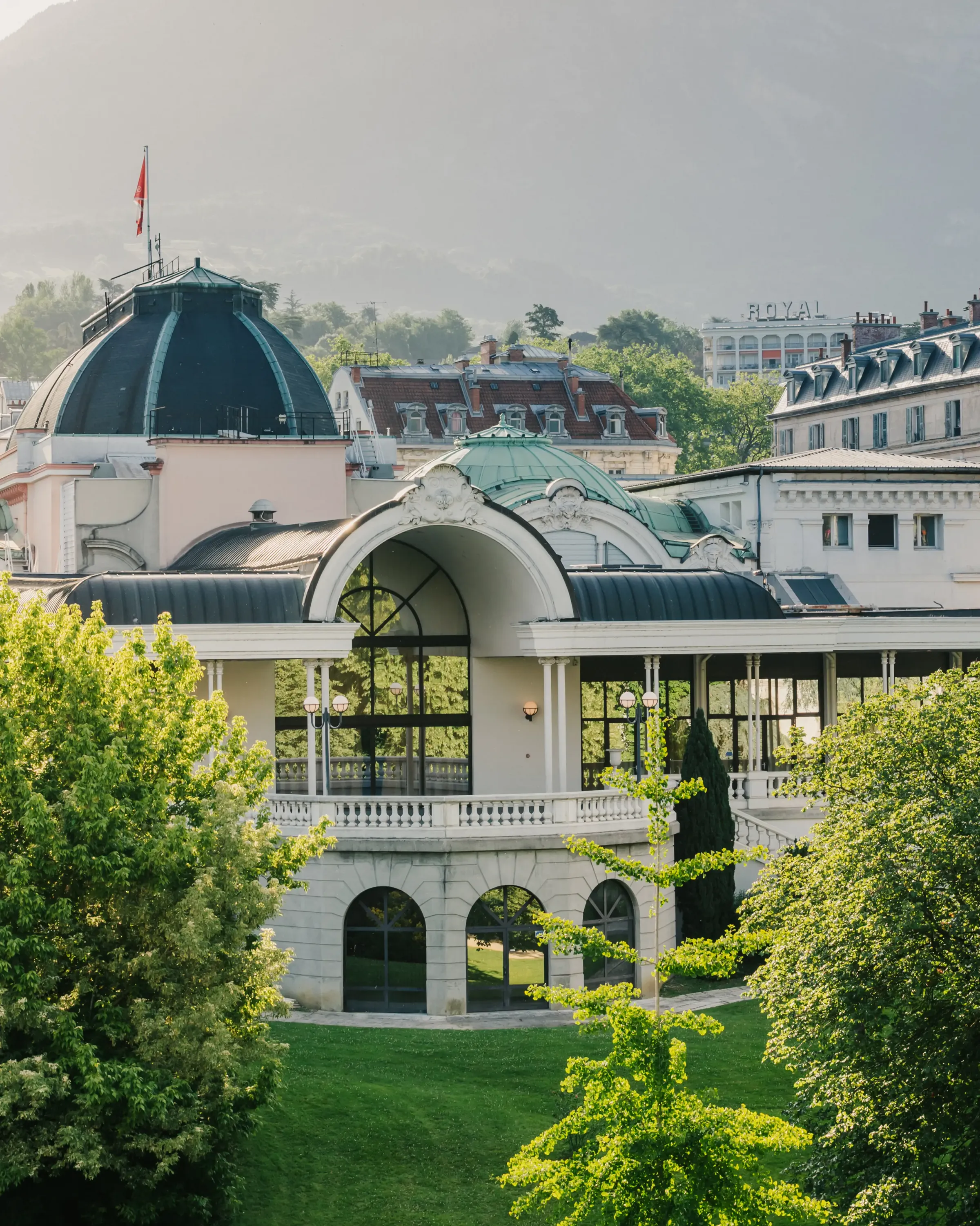 A building with classical architecture and a large skylight, surrounded by trees and lawns, with other structures and a flag in the background.