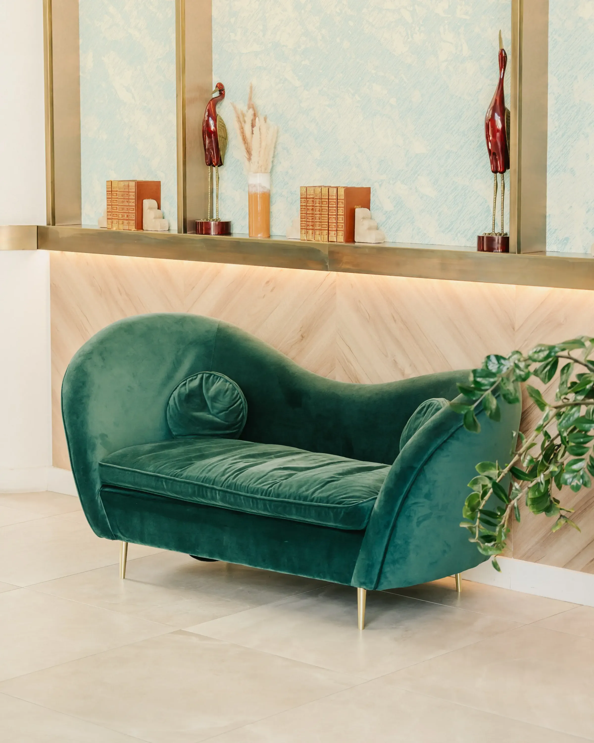 Ein grünes Samtsofa mit goldenen Füßen steht an einer hellen Holzwand unter einem Regal, das mit Gegenständen und Büchern dekoriert ist.