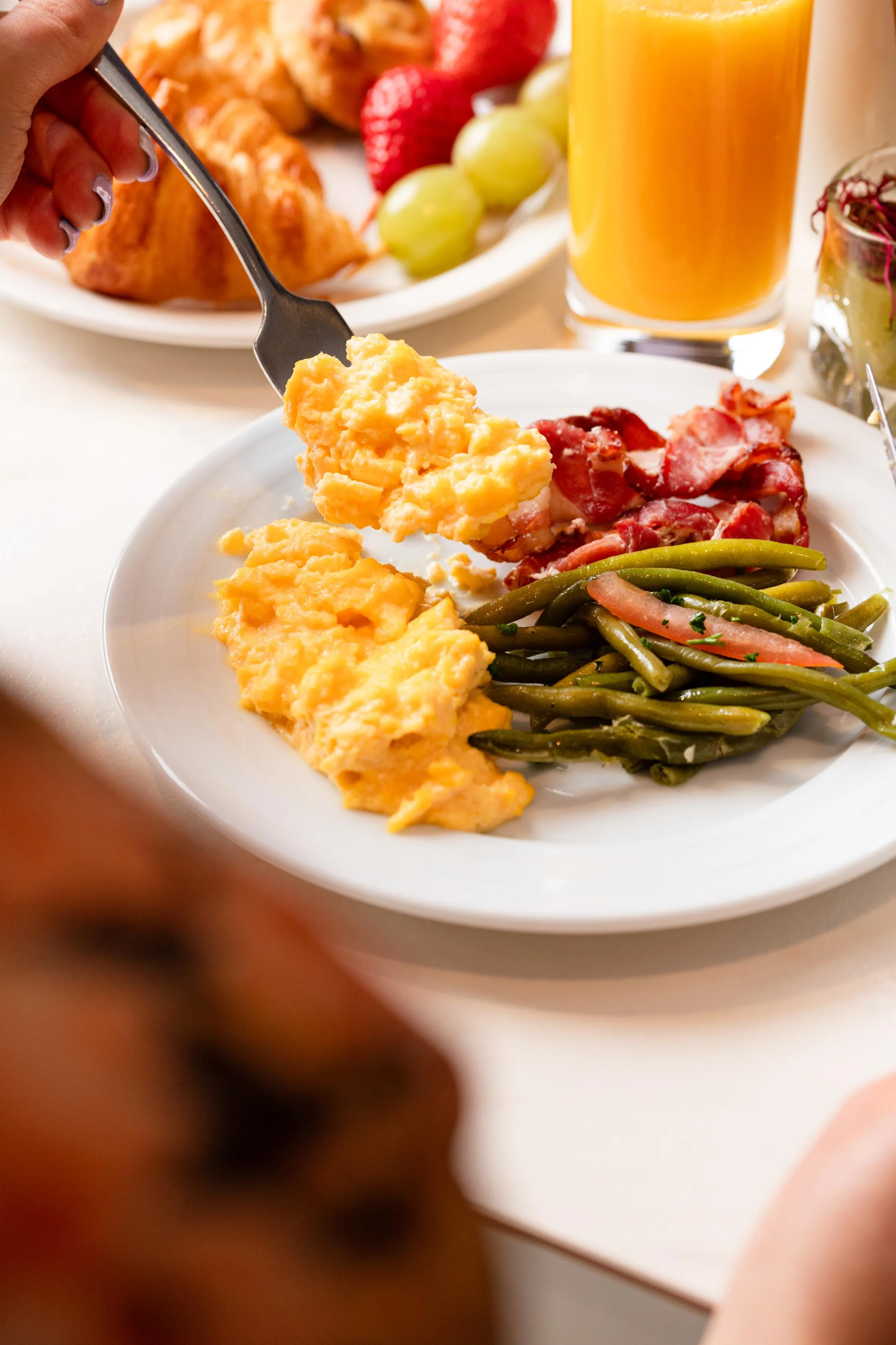 Une fourchette soulève une bouchée d'omelette sur une assiette contenant également des haricots verts, du bacon et des tomates, avec un verre de jus d'orange en arrière-plan