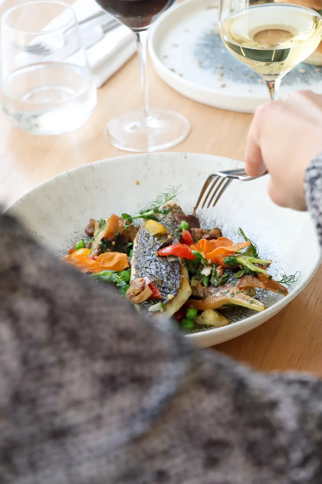 A grilled fish served with vegetables and carrots, on a white plate, with a hand holding a fork and wine glasses in the background.
