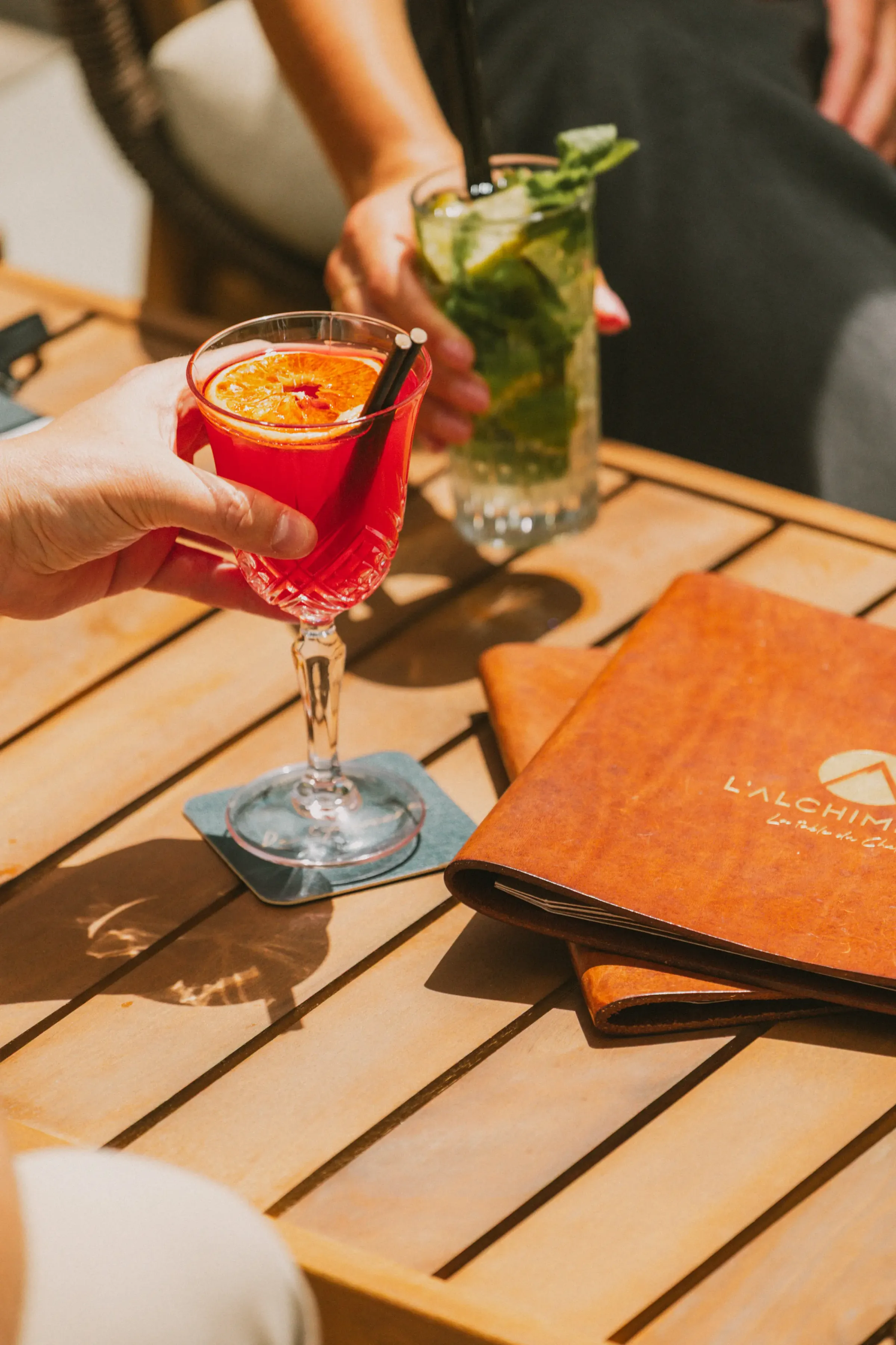 A hand holds a red cocktail glass with an orange slice, on a wooden table with menus and a water glass.