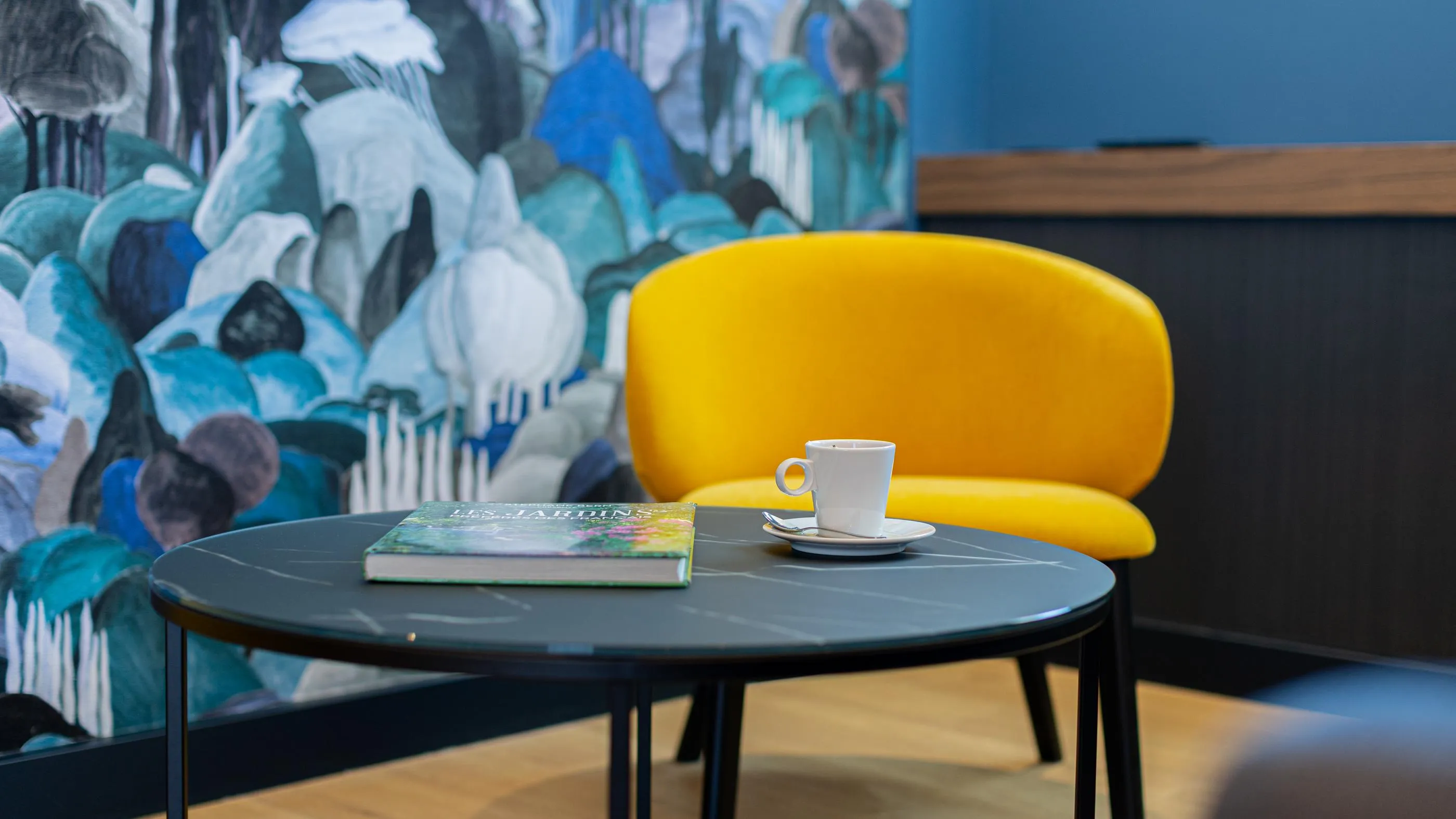 Une chaise jaune, un livre et une tasse de café sur une table ronde devant un mur décoré de motifs floraux colorés