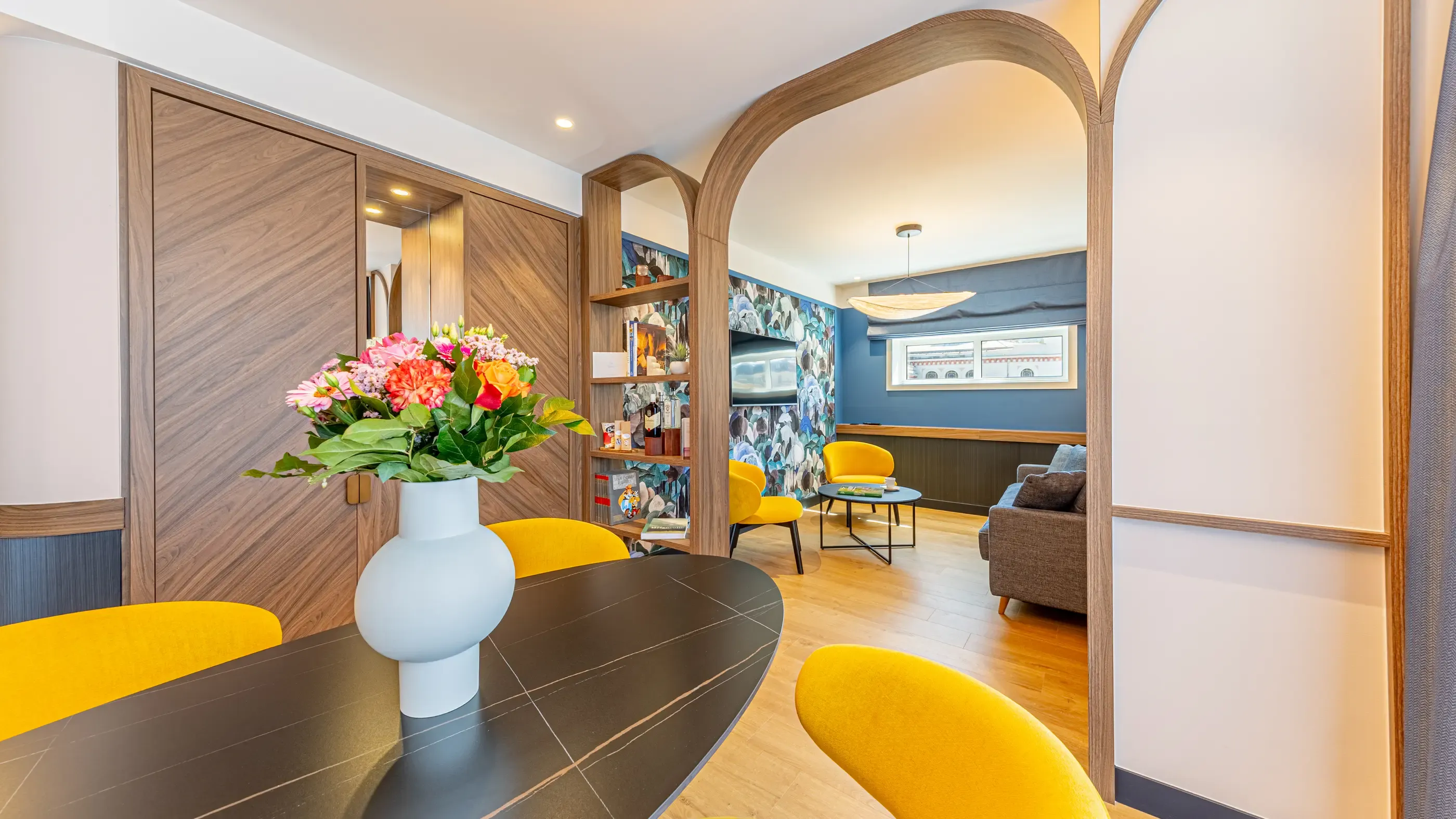 Modern living room with black table, yellow chairs, flower vase, and library in an adjacent room