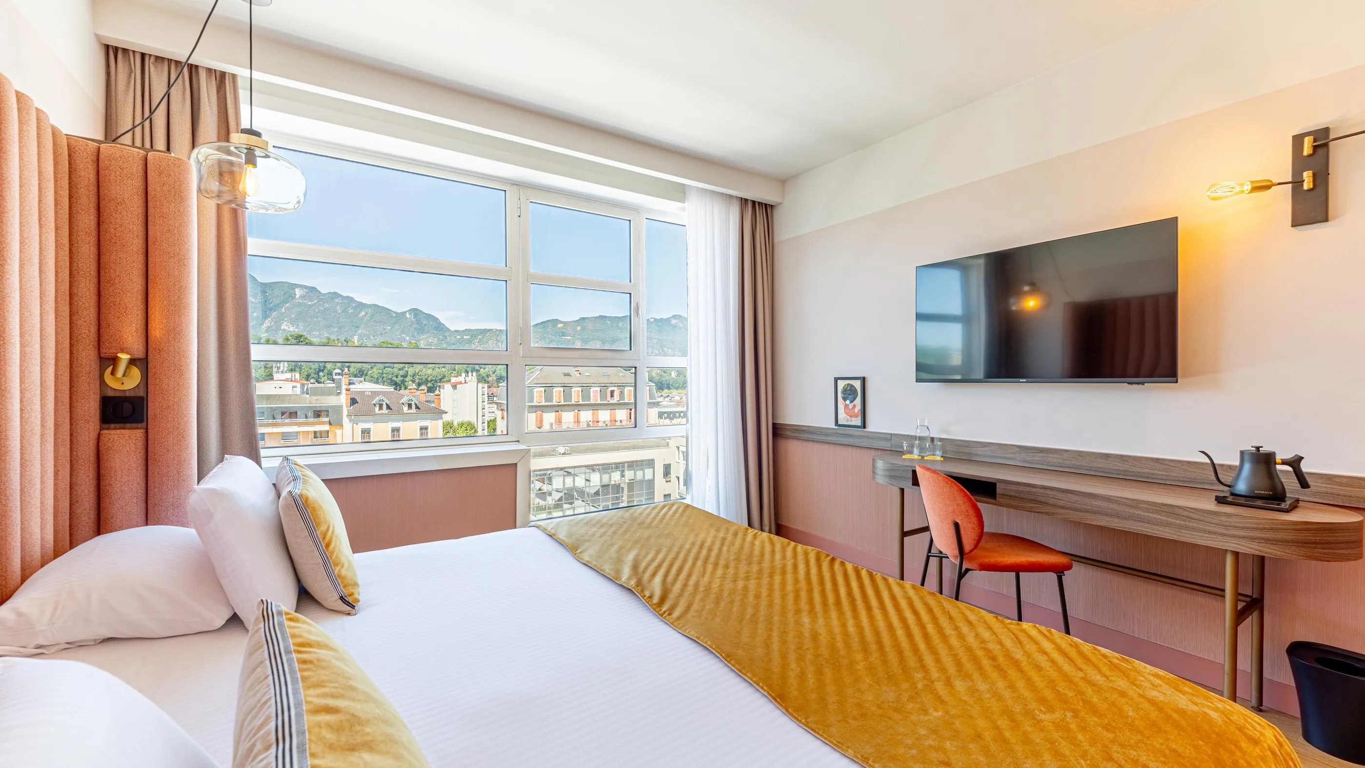 Chambre d'hôtel avec deux lits, une fenêtre offrant une vue sur des montagnes et des bâtiments, un bureau avec une chaise et un téléviseur mural