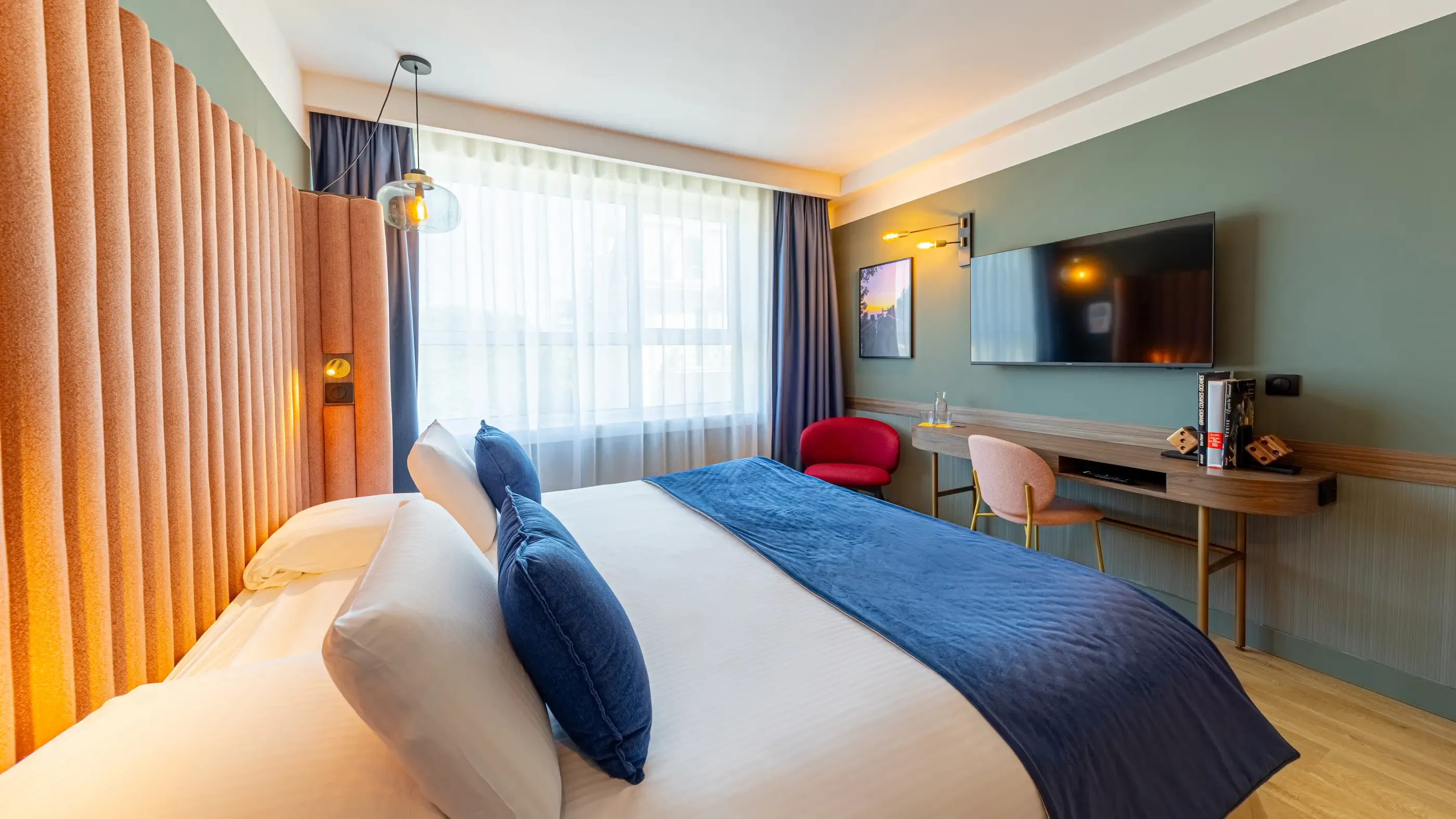 Hotel room with a double bed, a television, a desk, a chair, and a large window with curtains.