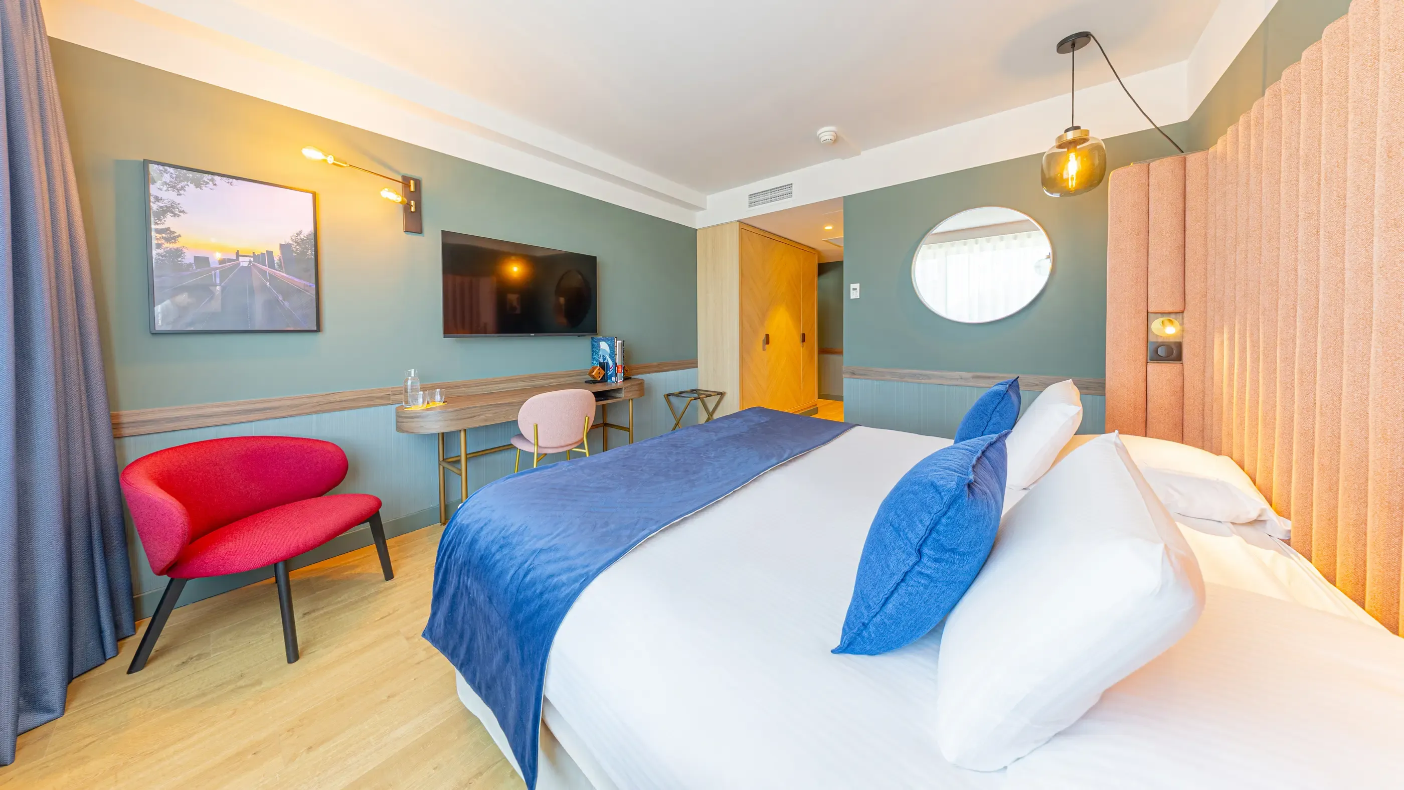 Chambre d'hôtel avec deux lits, un fauteuil rouge, une table avec une chaise, deux écrans muraux et une porte ouverte
